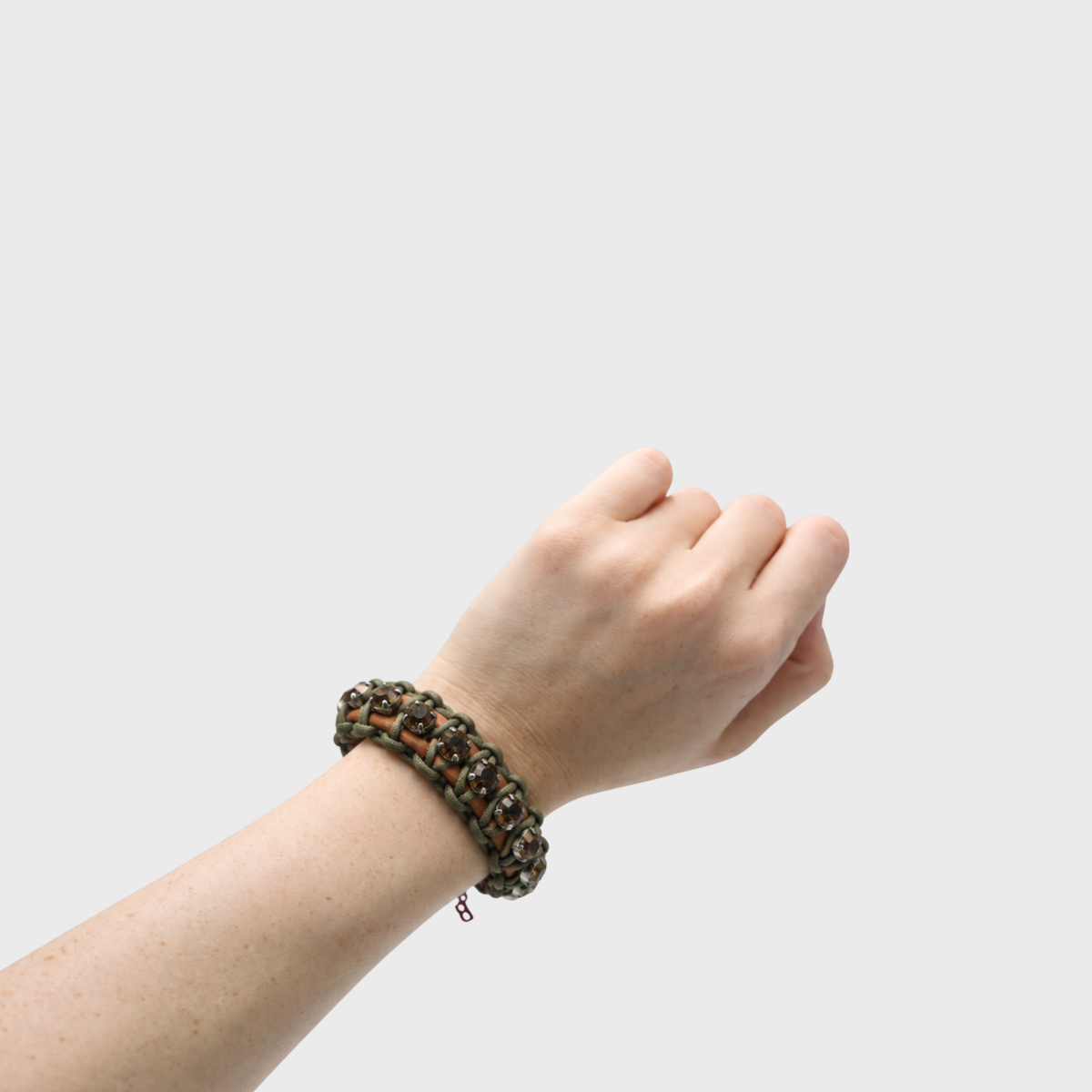 A close-up of a person's wrist wearing a multi-strand woven macrame bracelet in dark brown and olive green with brown stones, set against a solid light gray background.