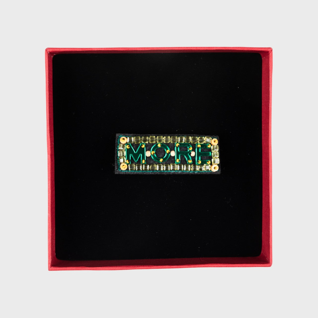 An embroidered green brooch with MORE written in a box.