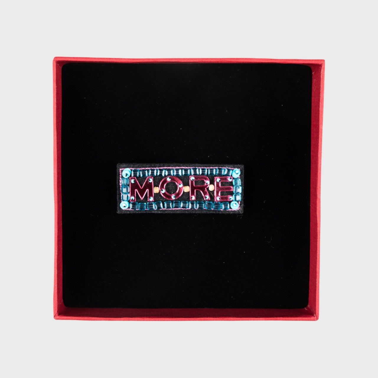 An embroidered brooch with MORE written in pink in a box.