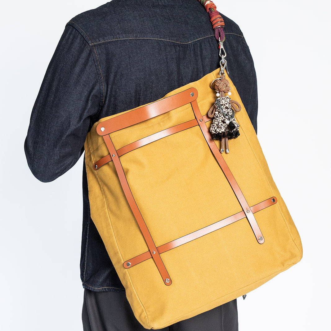 A mustard yellow canvas tote bag with brown leather straps, worn crossbody by a person wearing a dark denim shirt and dark pants, shown from the back against a white studio background.