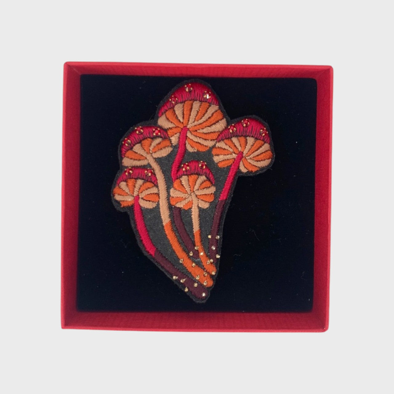 An embroidered brooch representing a bucket of red mushrooms with red, orange and beige details in a box.