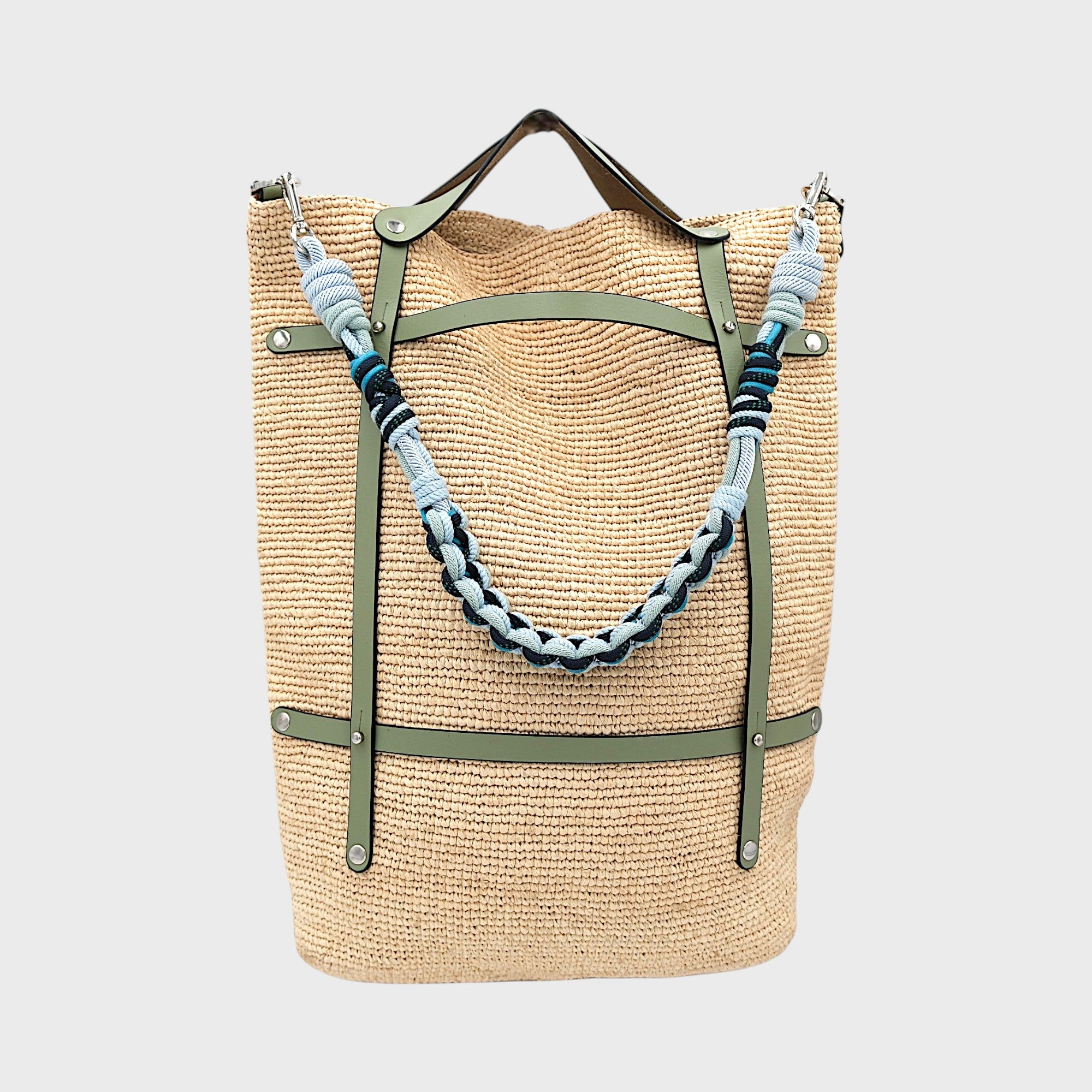 A raffia tote bag in a natural color, featuring green leather straps and a decorative beaded shoulder strap with dark and light blue details.