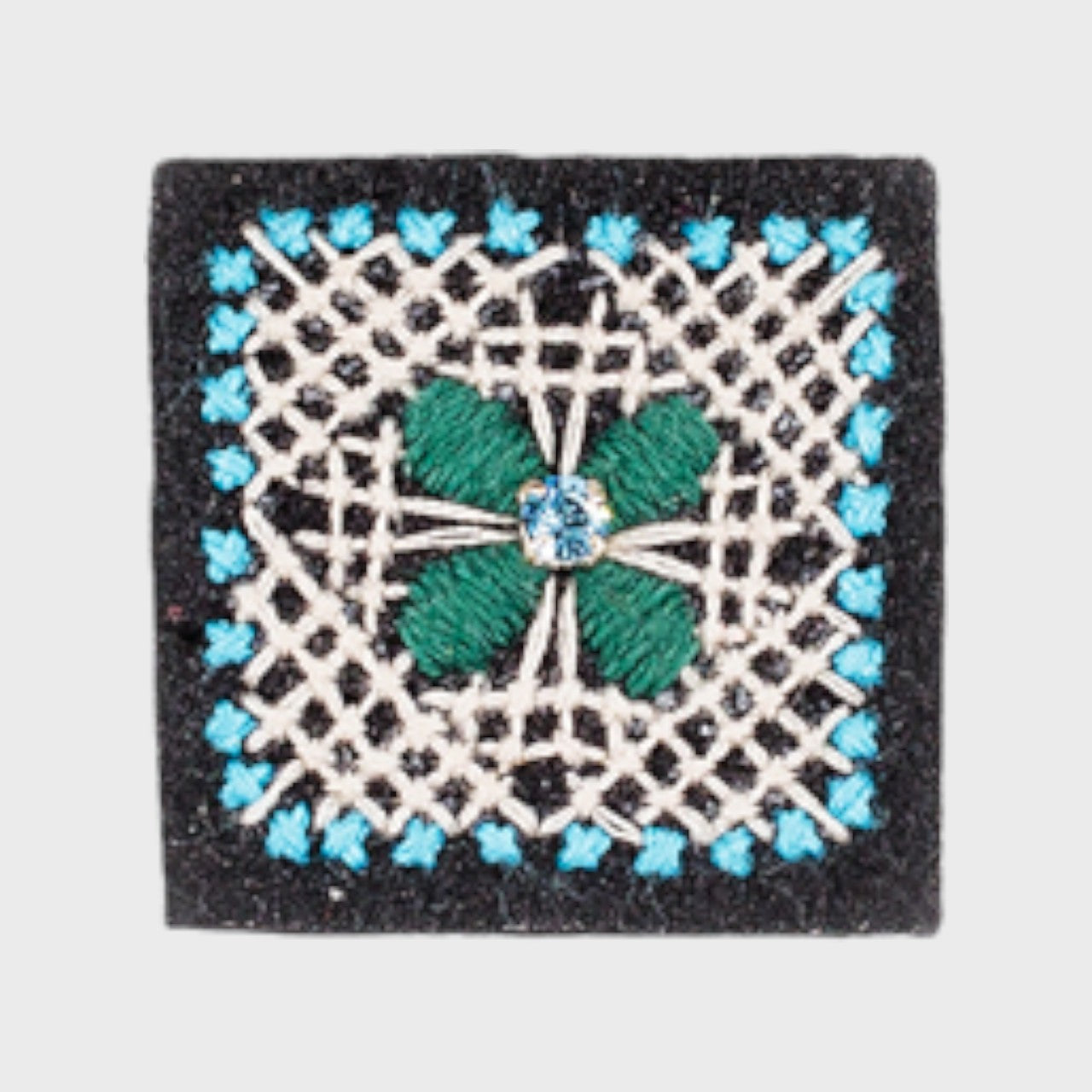 An embroidered brooch representing a square with blue and white details and a green lower in the middle.
