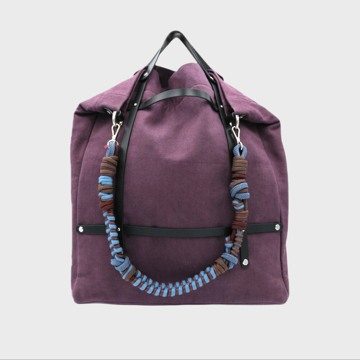 Purple tote bag with black leather straps and a unique macrame shoulder strap design on a light gray background.