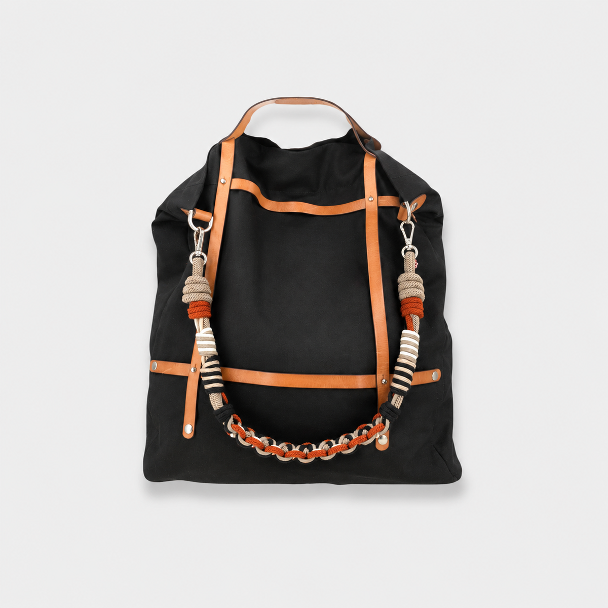 A black canvas tote bag with tan leather straps and handles, decorated with woven macrame shoulder strap shown against a light gray background.