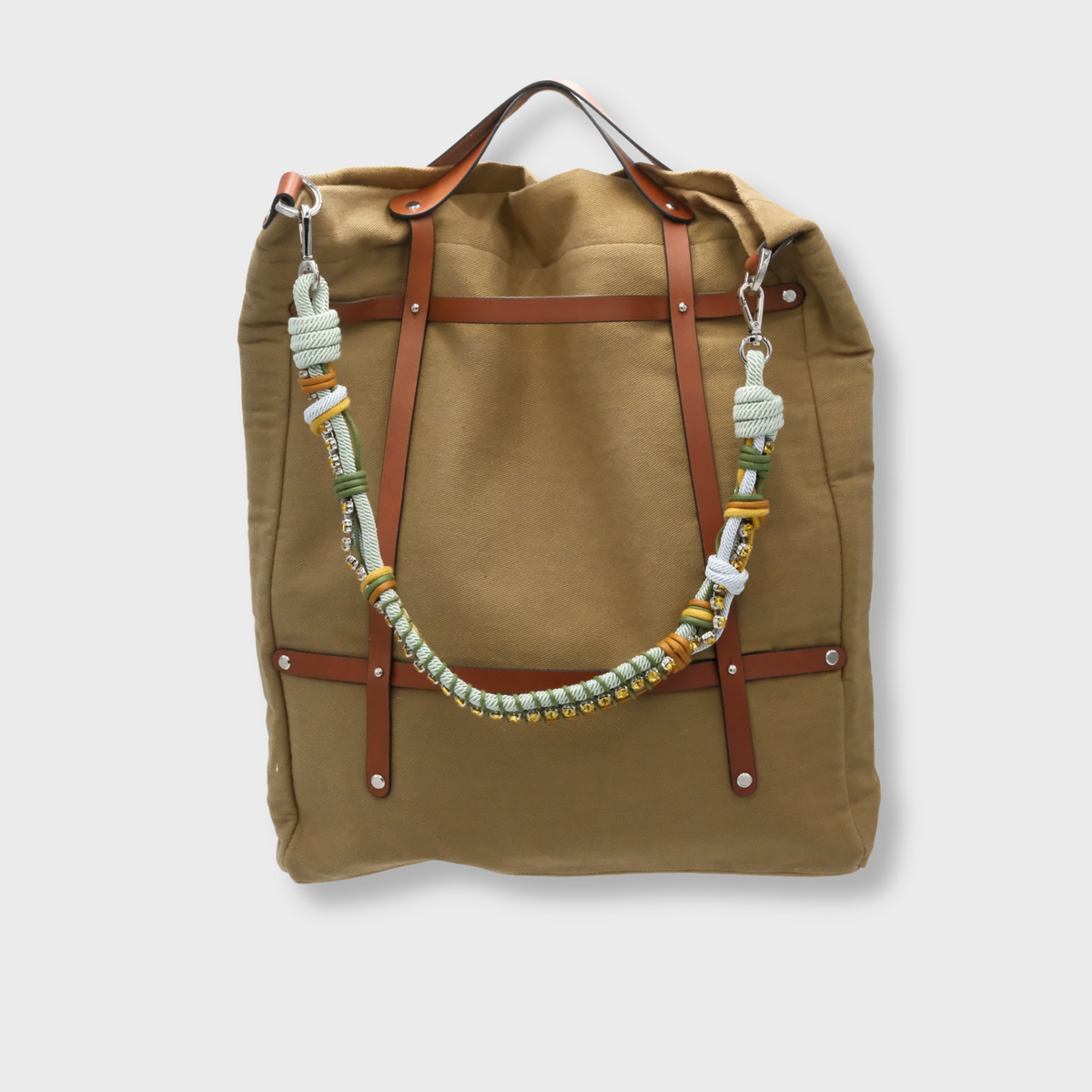 A canvas tote bag in a Taupe color, featuring brown leather straps and a decorative beaded shoulder strap with green, white, and gold details.