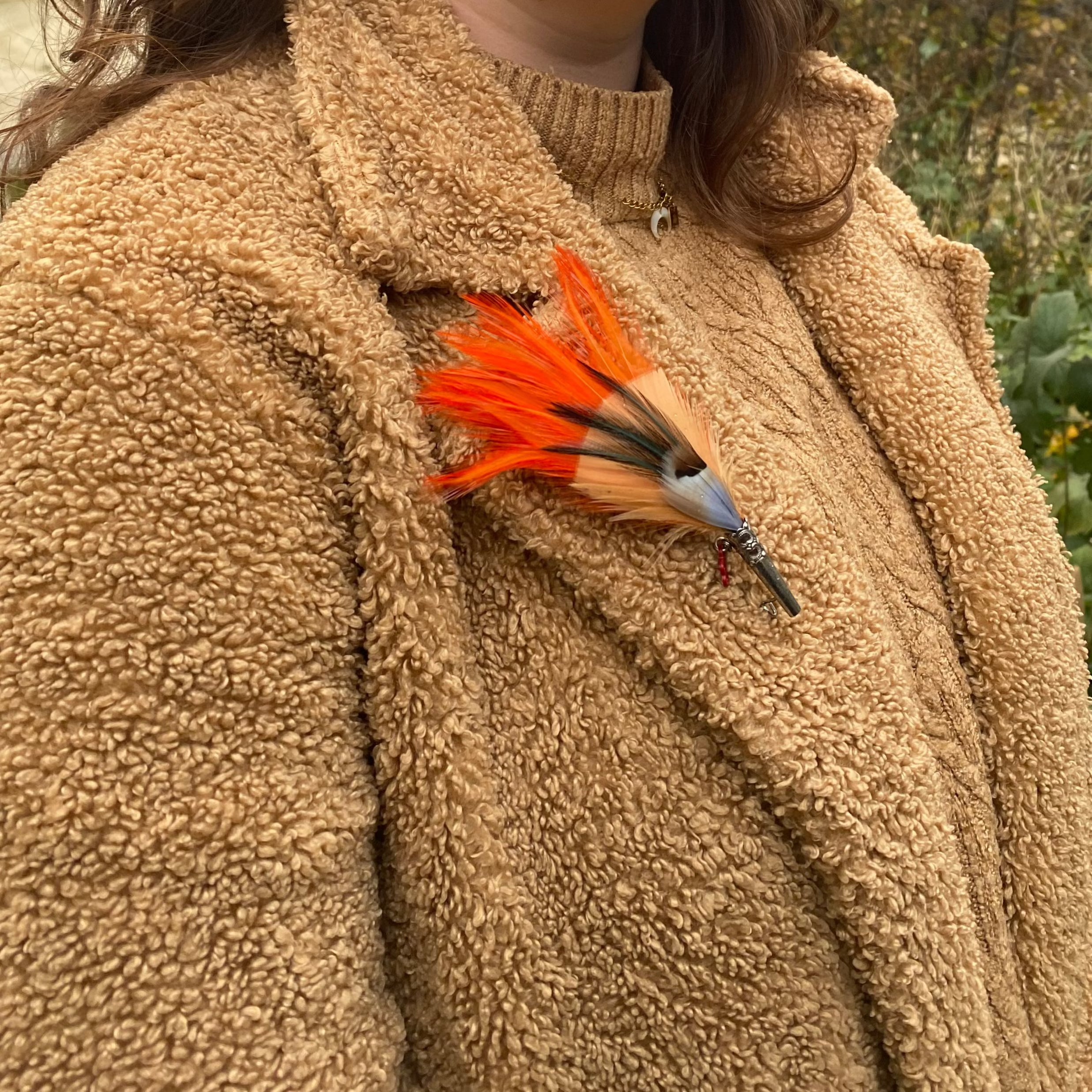 A person wearing a jacket with a dyed orange feather brooch pinning it.