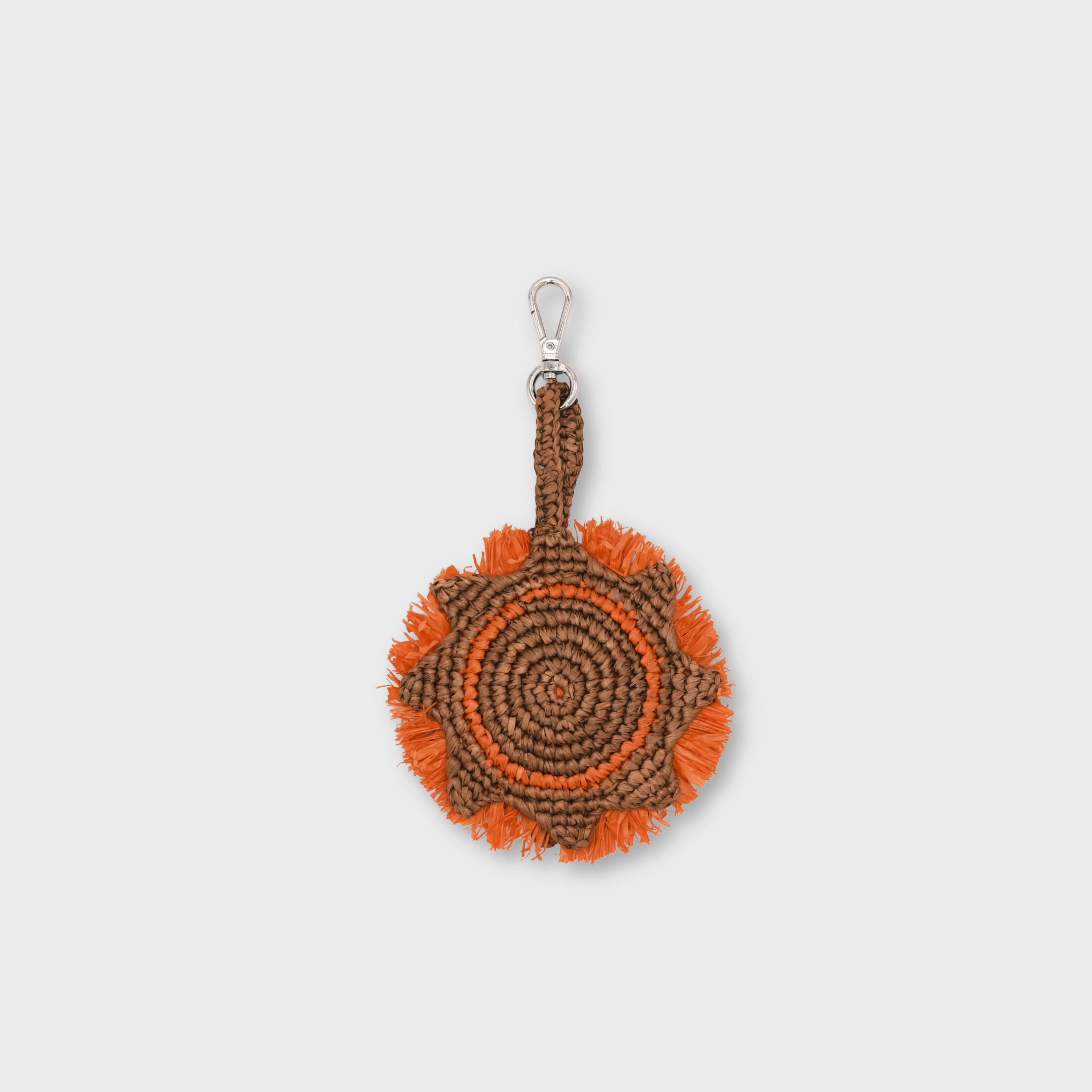 A circular raffia keychain with a spiral pattern in brown and orange, representing a sun and featuring fringe details and a silver metal lobster clasp at the top.