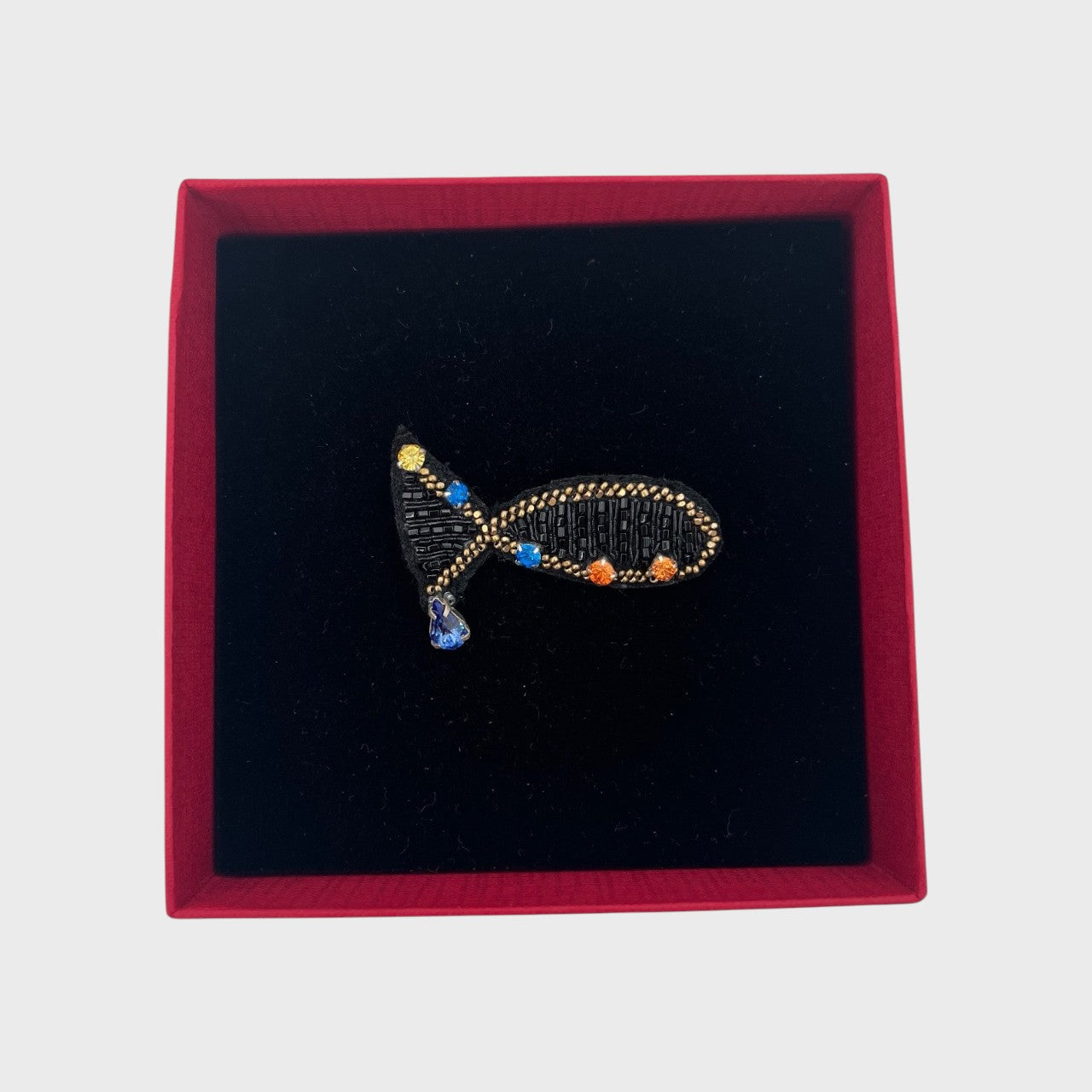 An embroidered brooch representing a small black ish with golden details in a box.