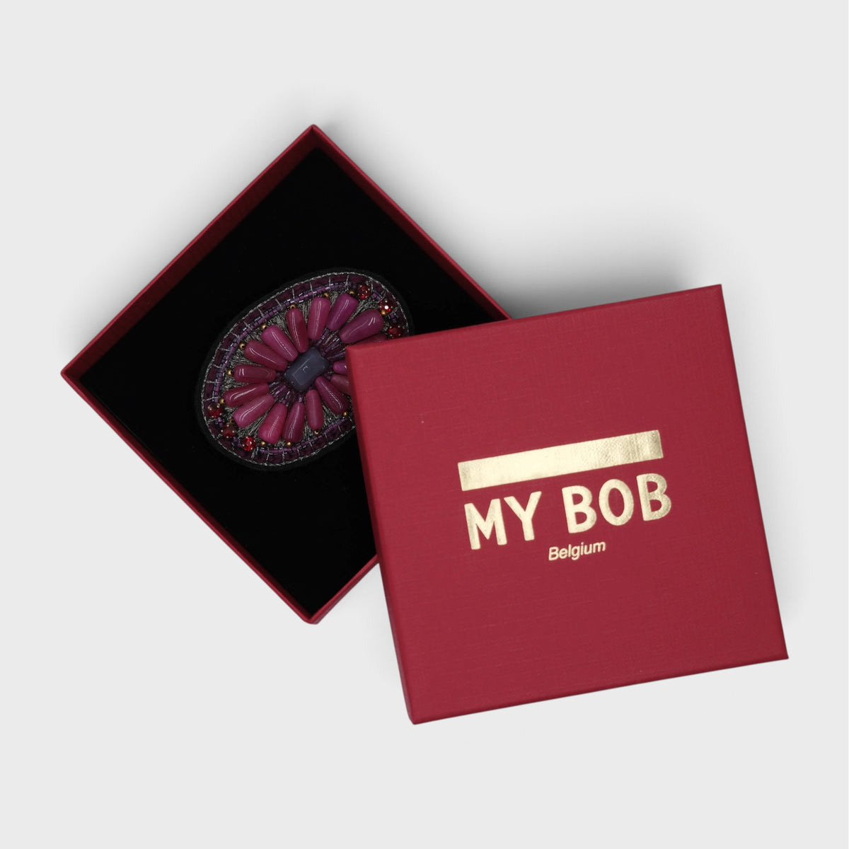 An oval embroidered brooch with a pink and purple floral design, displayed inside a red presentation box with a black velvet insert.