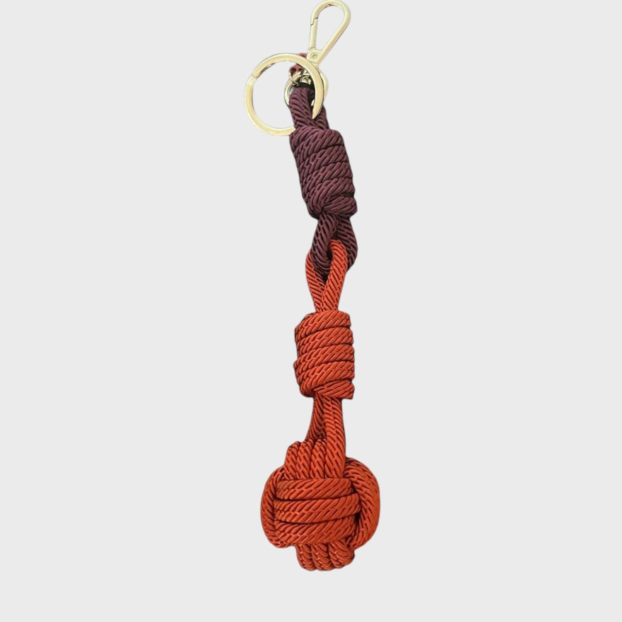 A macramé keychain representing a ball inspired by sailors knots. The straight upper part is dark purple and the ball part is dark orange.