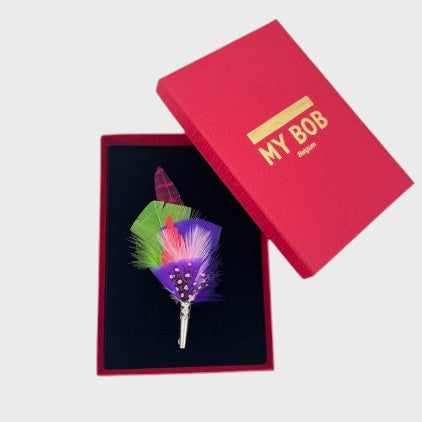 Purple, green and pink feather brooch in a box on a light gray background
