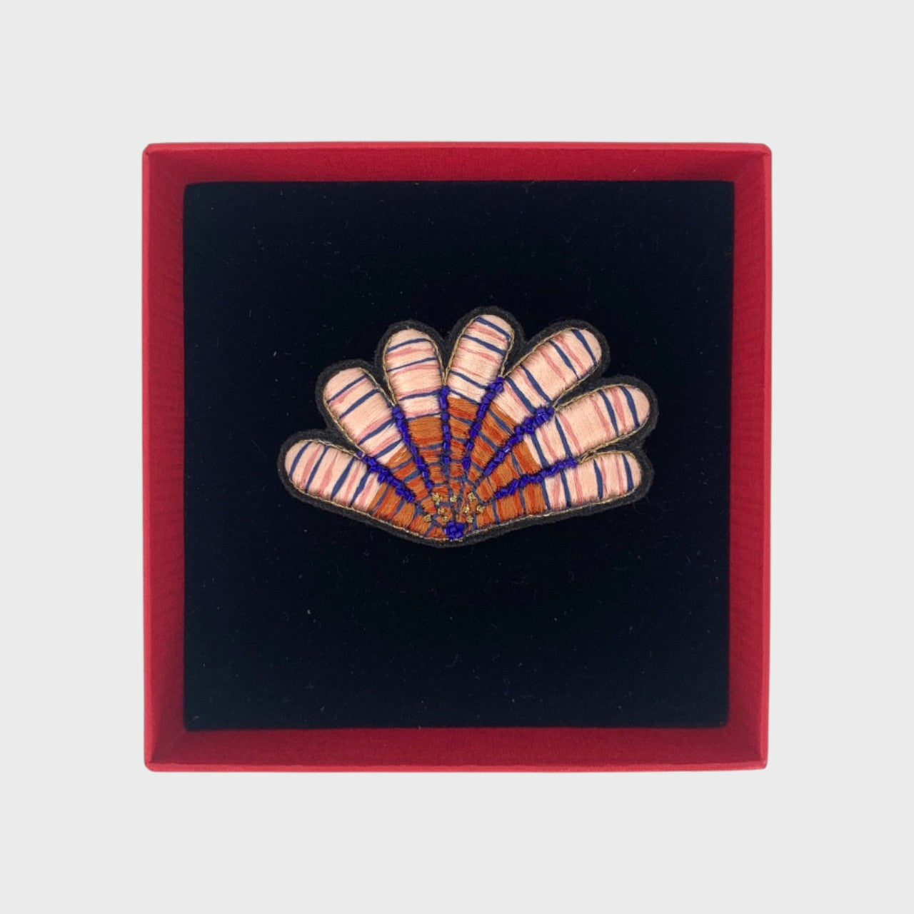 An embroidered pink shell brooch with blue, pink and brown details in a box.