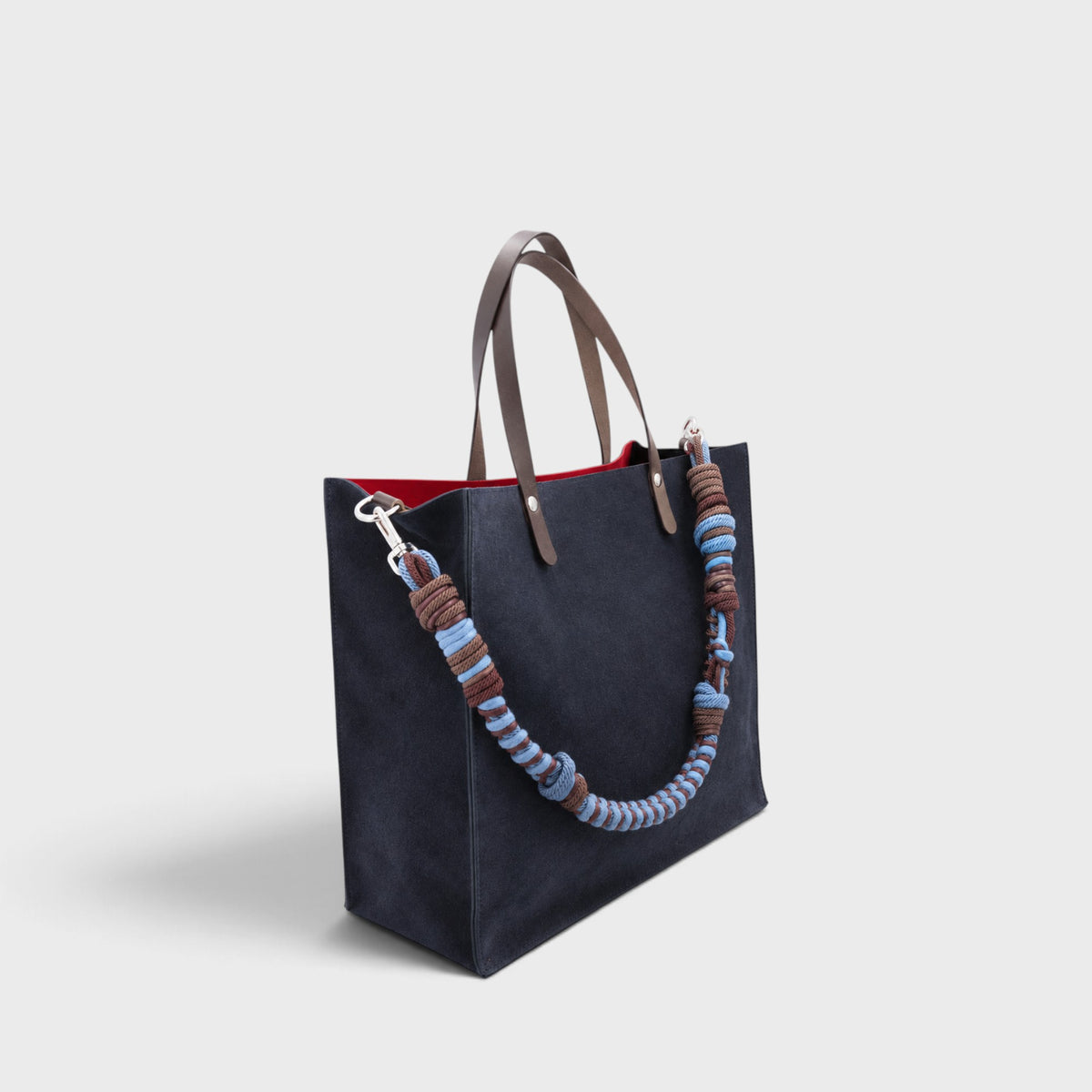 A large dark navy blue suede cabas bag with brown leather handles and a long woven rope shoulder strap in brown, blue, and light blue, shown against a light gray background.