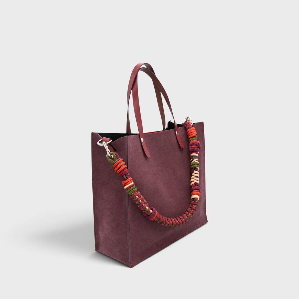 A large structured tote bag in deep burgundy suede, featuring dual leather handles and a thick, colorful braided shoulder macrame strap made in red, orange, green, and cream, shown against a light gray background.