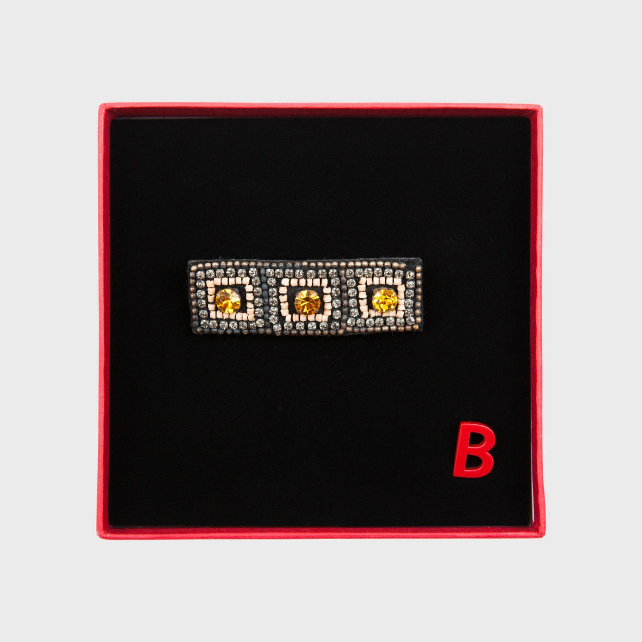 An embroidered brooch representing a silver tiara in a box.