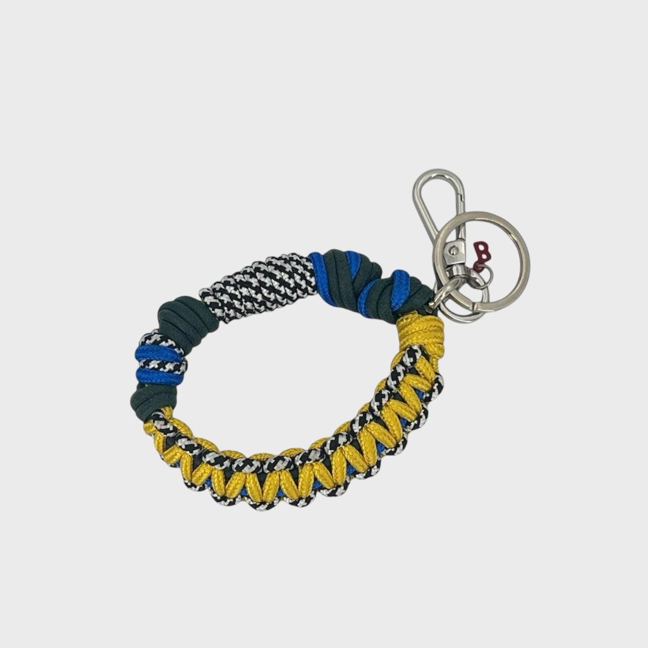 A woven macramé key chain with yellow, black and white, dark green and blue patterns, inspired by celtic knots, finished with silver-tone metal hardware.