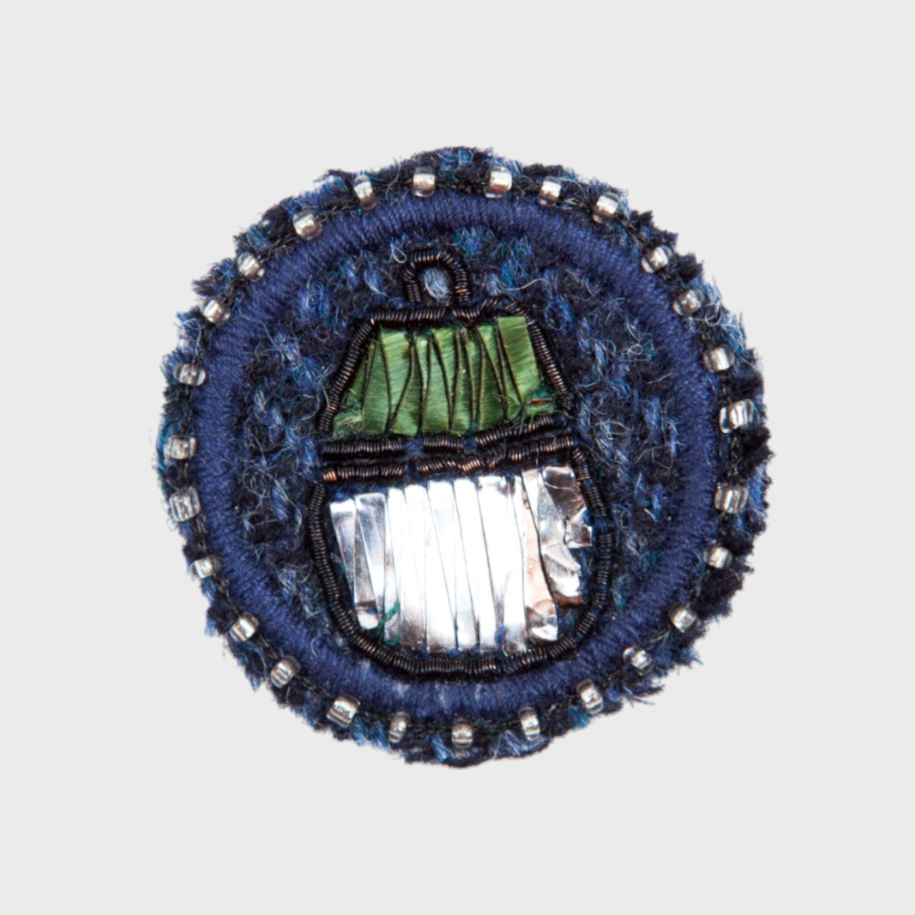 An embroidered brooch representing a green and silver acorn in the middle of a blue circle. 
