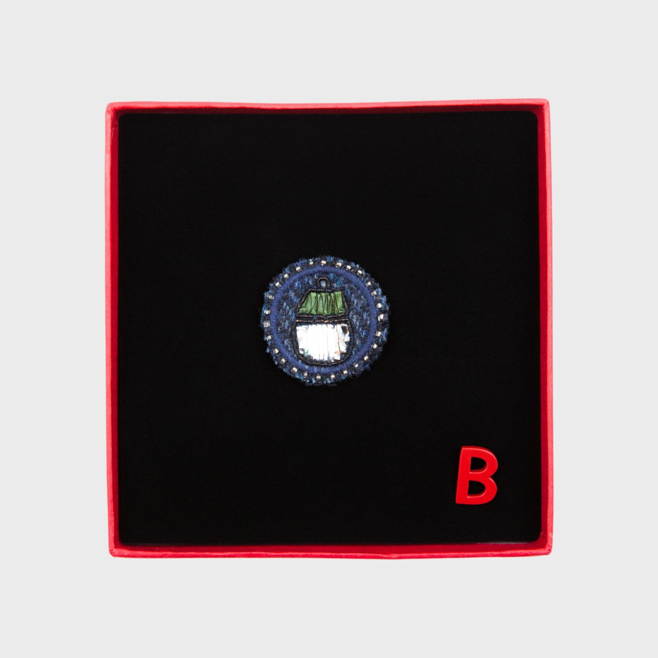 An embroidered brooch representing an acorn in the middle of a blue circle in a box. 