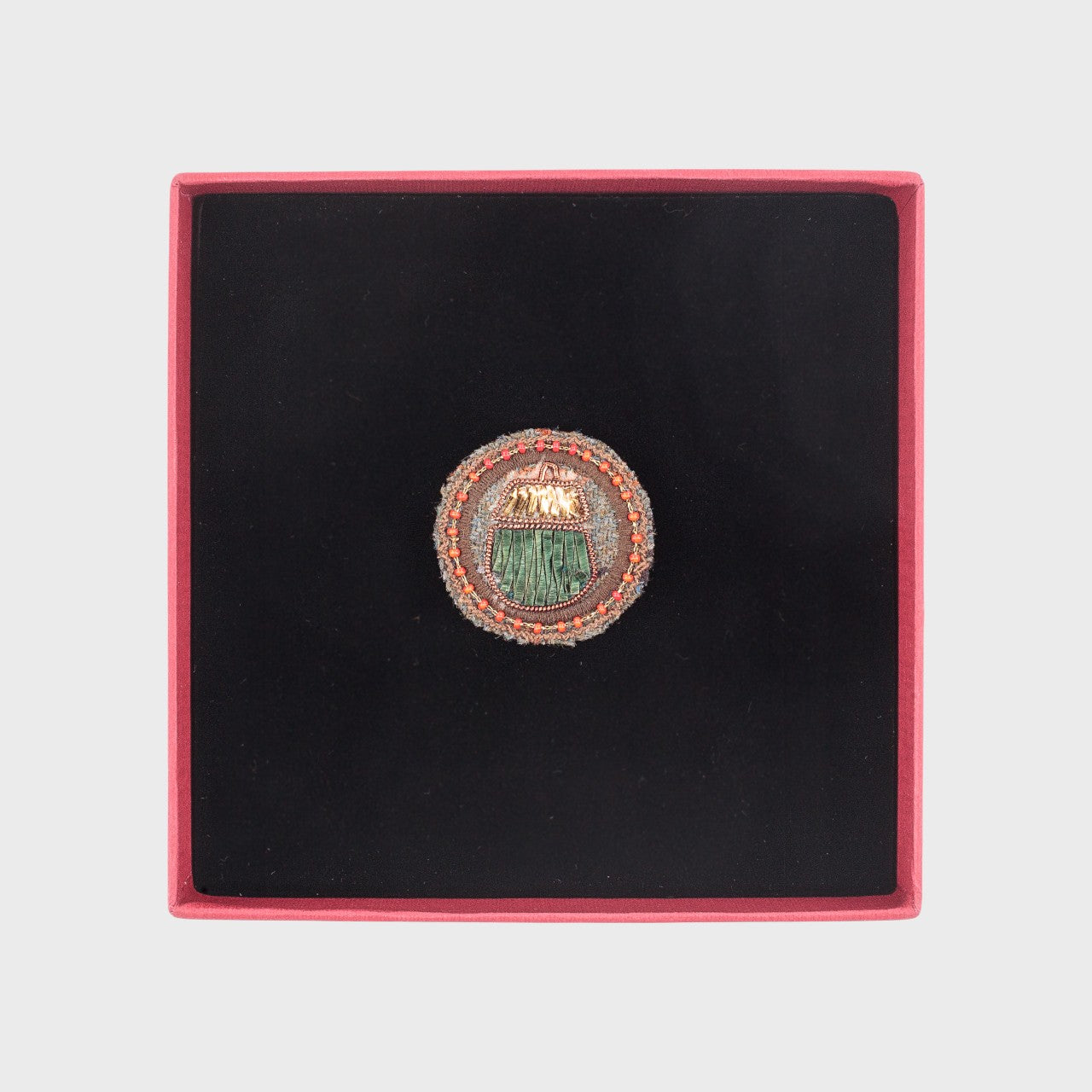 An embroidered brooch representing an acorn in the middle of a brown circle in a box. 