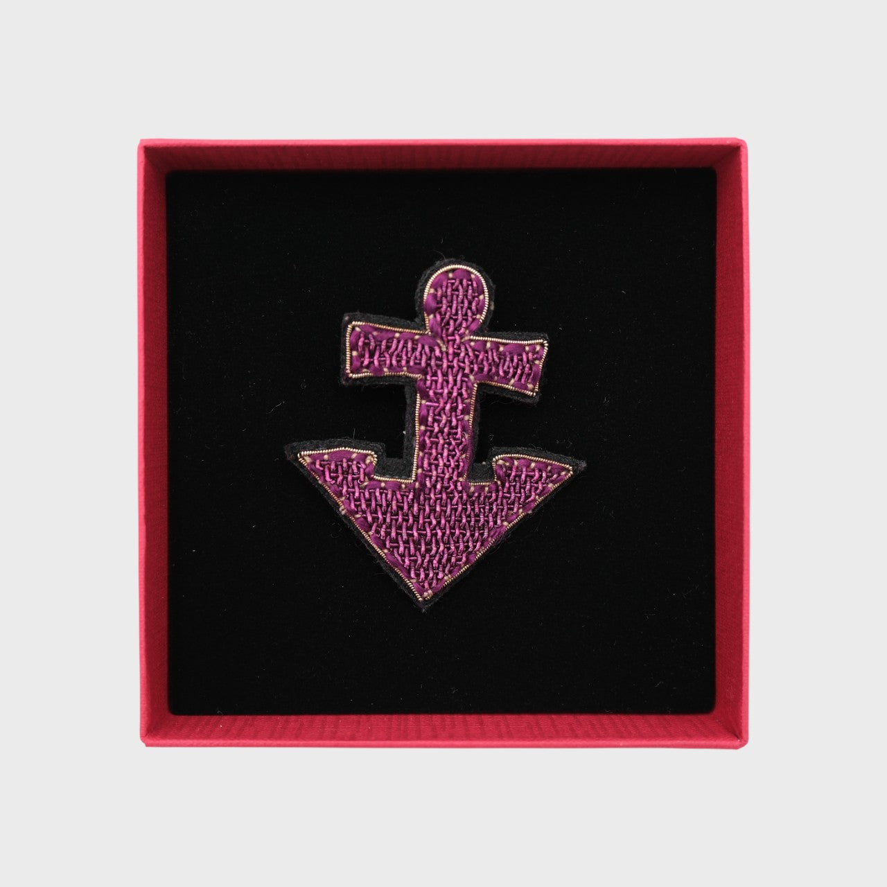 An embroidered brooch representing a purple anchor in a box. 