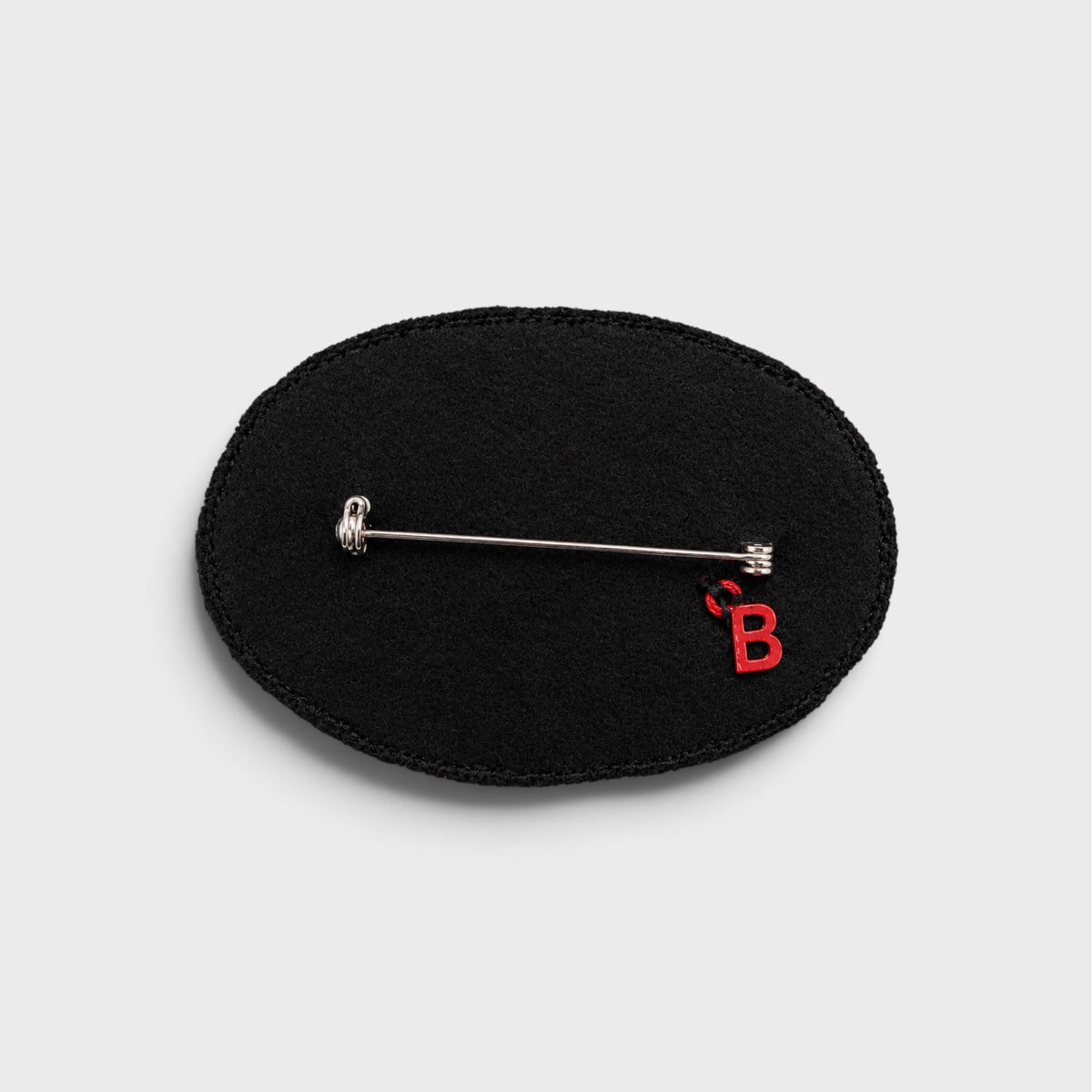 A black oval fabric pin brooch with a silver safety pin clasp and a small red embroidered letter 'B' on the bottom right, shown on a light gray background.