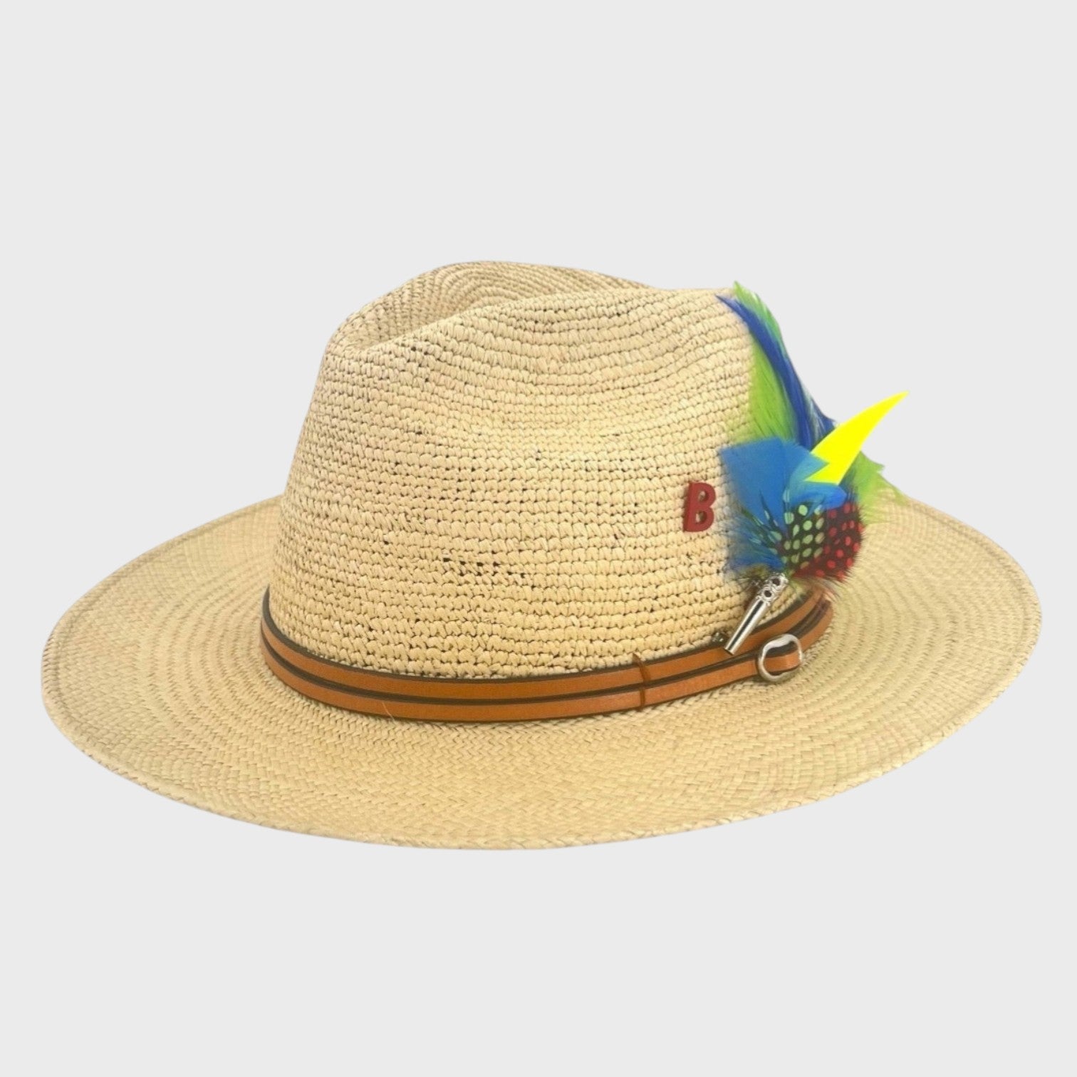 A beige raffia hat with a brown leather band around it and a blue, green and yellow feather brooch pinned on it. 
