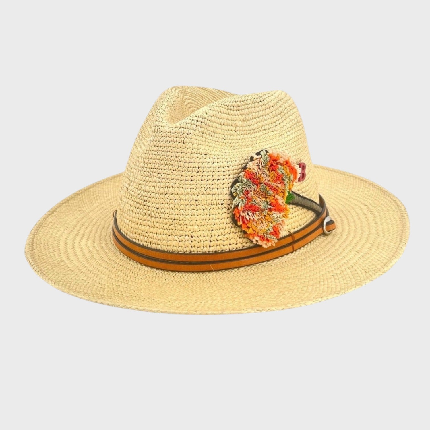 A beige raffia hat with a slim brown leather band around it and an orange embroidered carnation flower brooch pinned on it. 