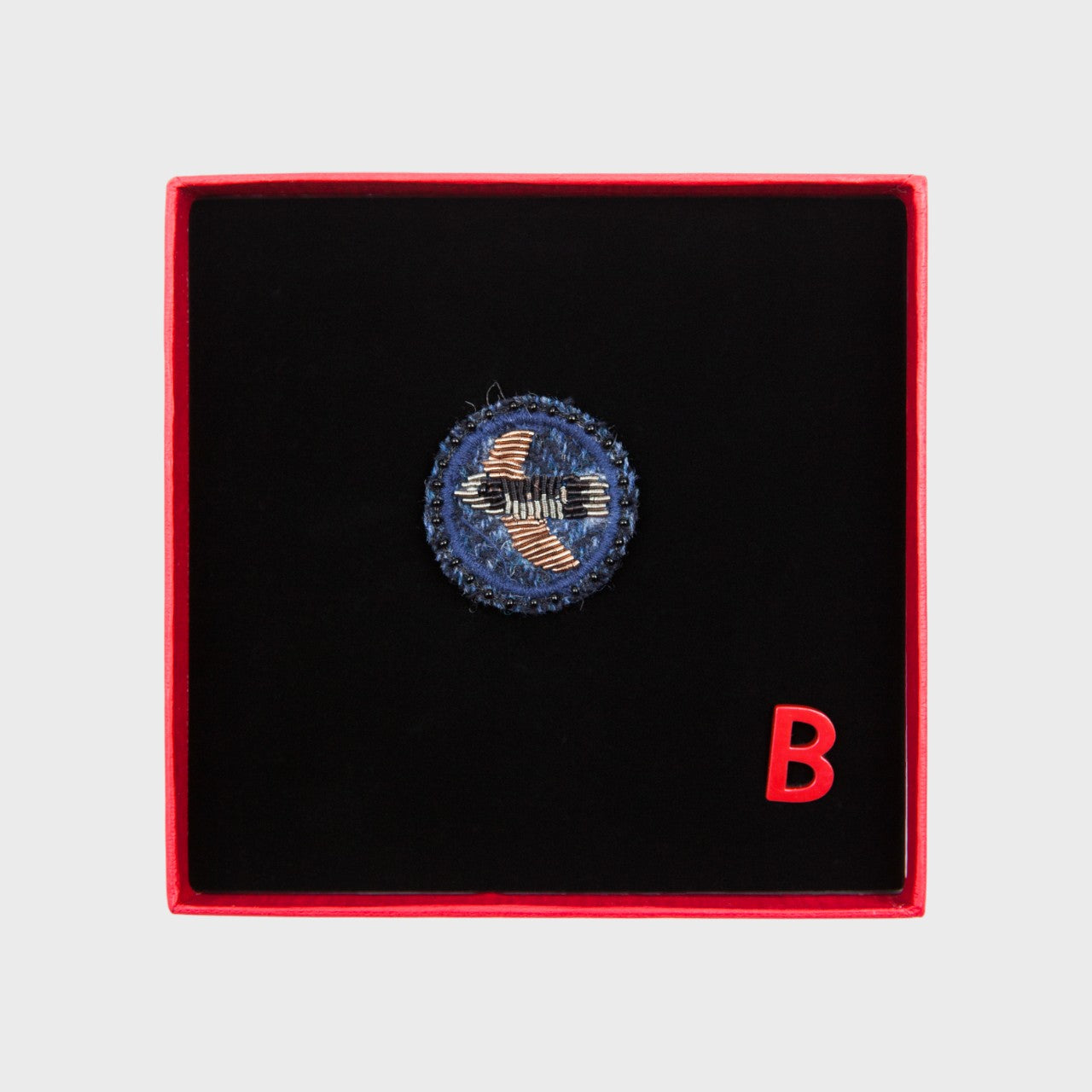 An embroidered brooch representing a bird in the middle of a blue circle in a box. 