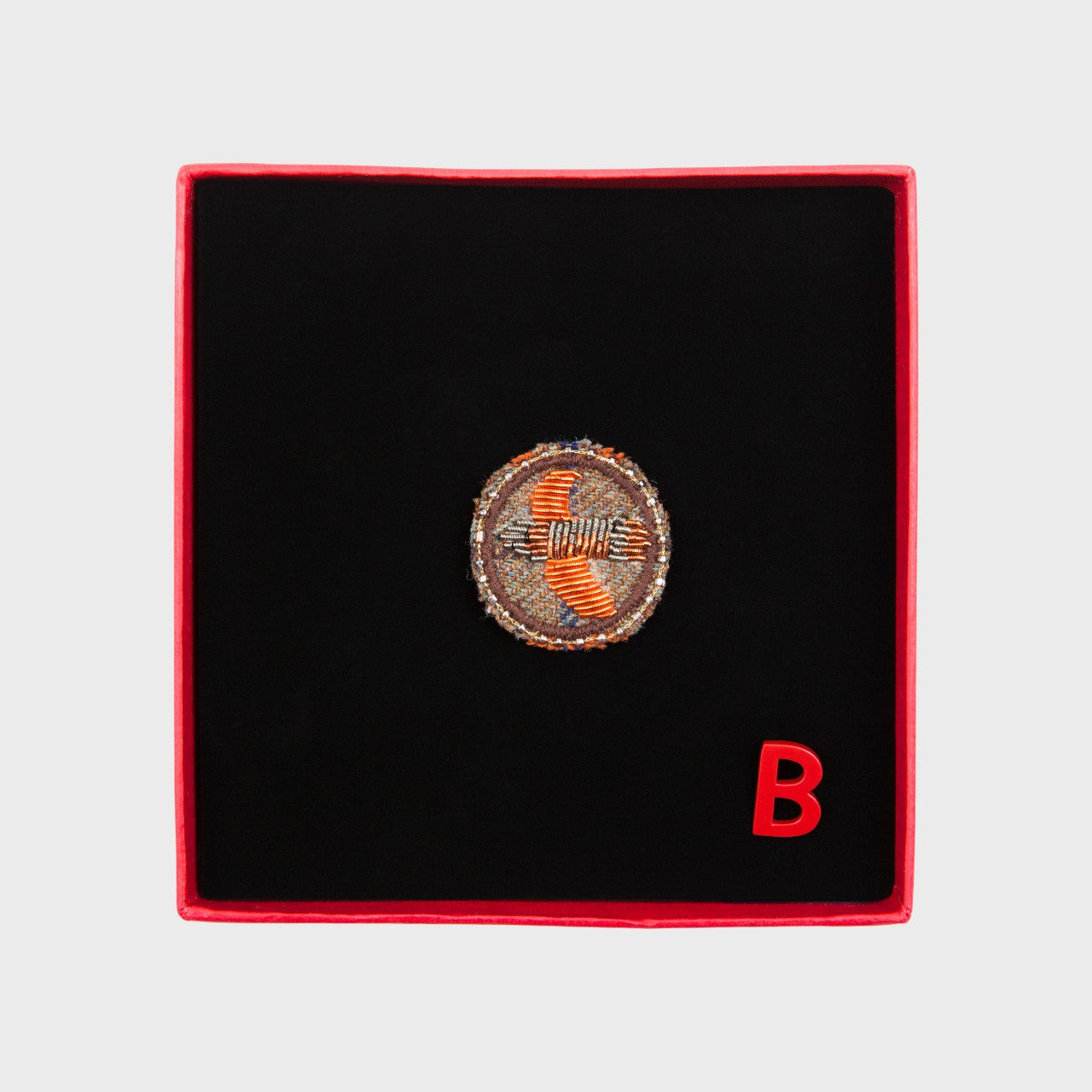 An embroidered brooch representing an orange bird in the middle of a circle in a box. 