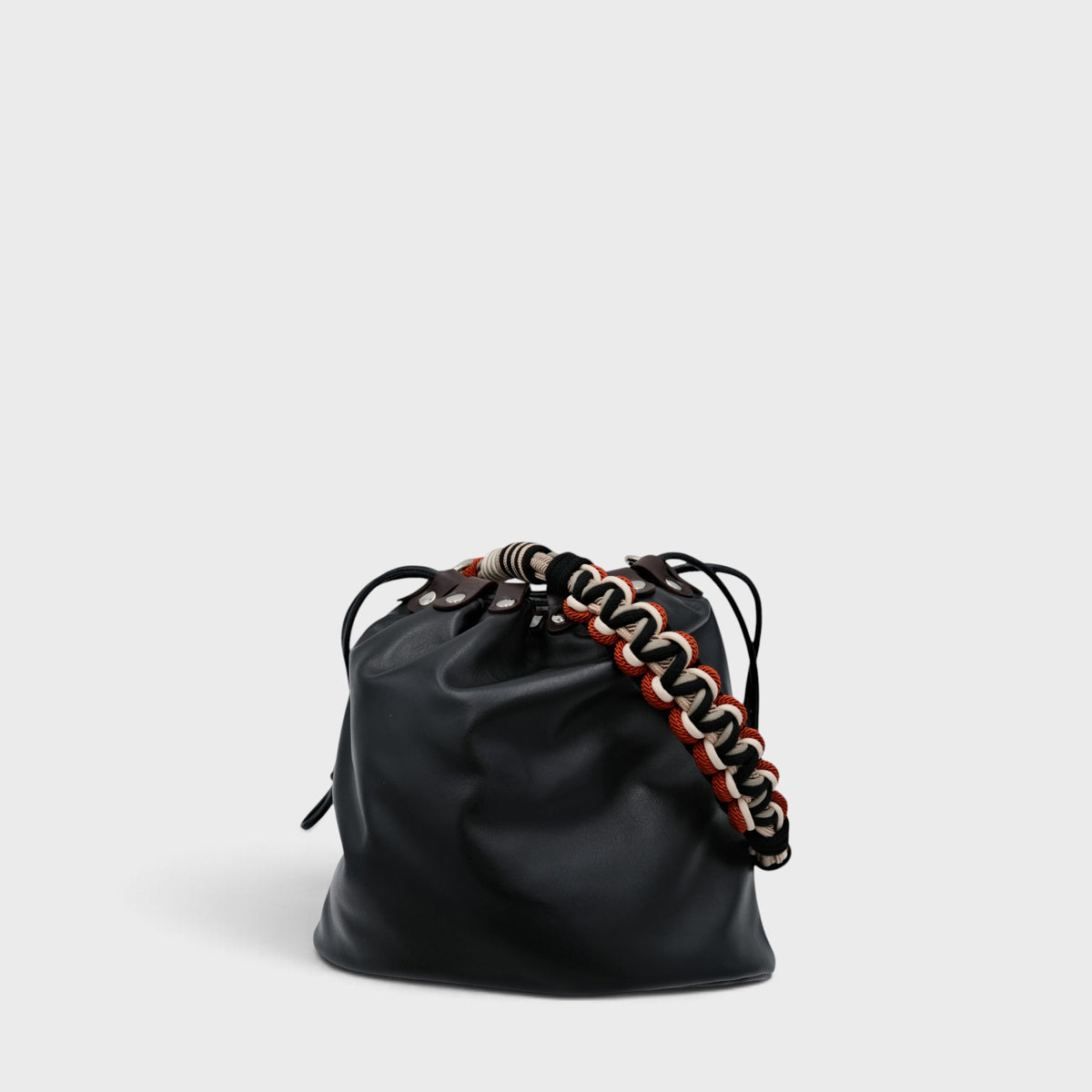 A black leather bucket bag with a chunky woven macrame strap in black, white, and red, shown on a light gray background.