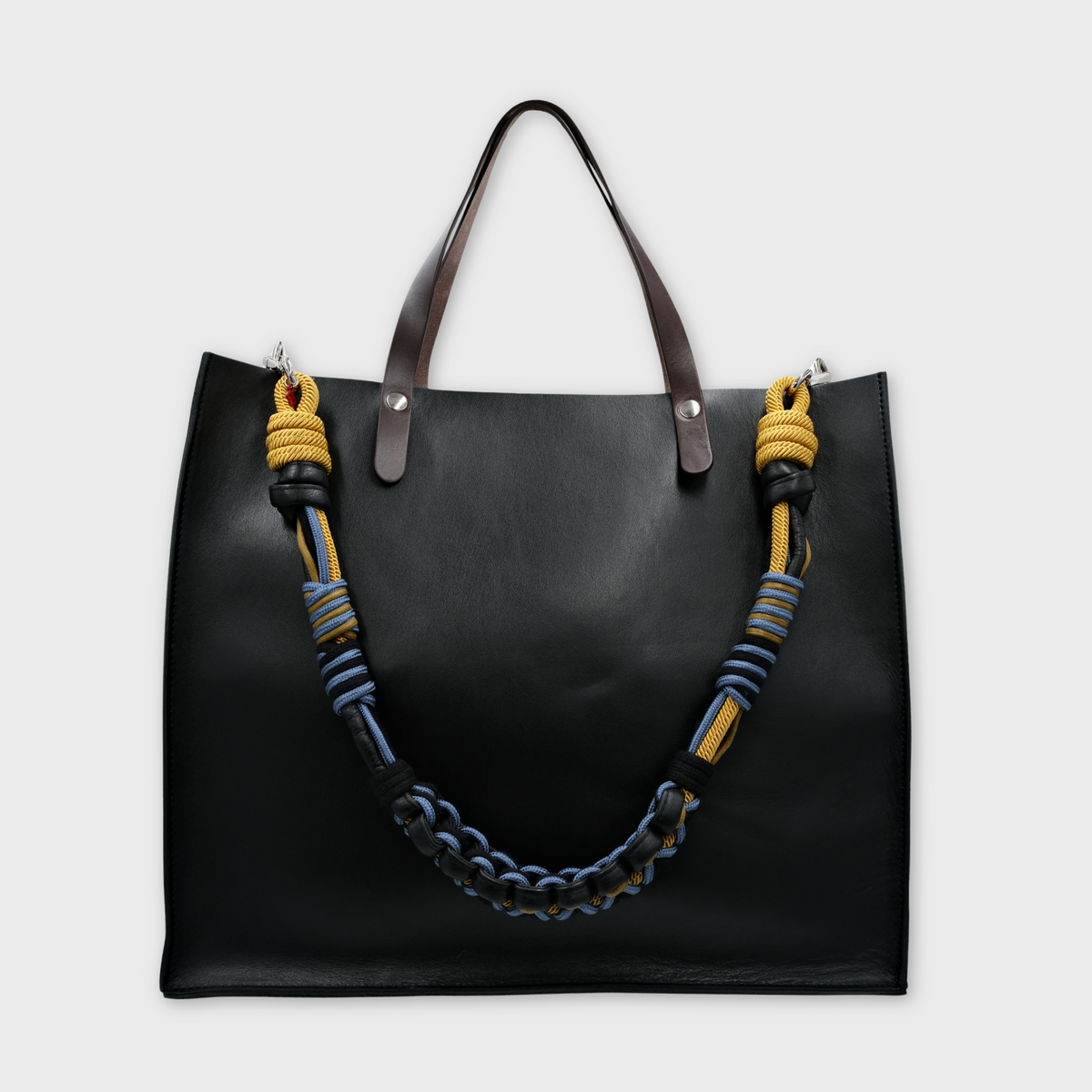 A black leather cabas bag with dark brown leather handles and a distinctive woven shoulder strap featuring yellow, blue, and black stripes, set against a light gray background.