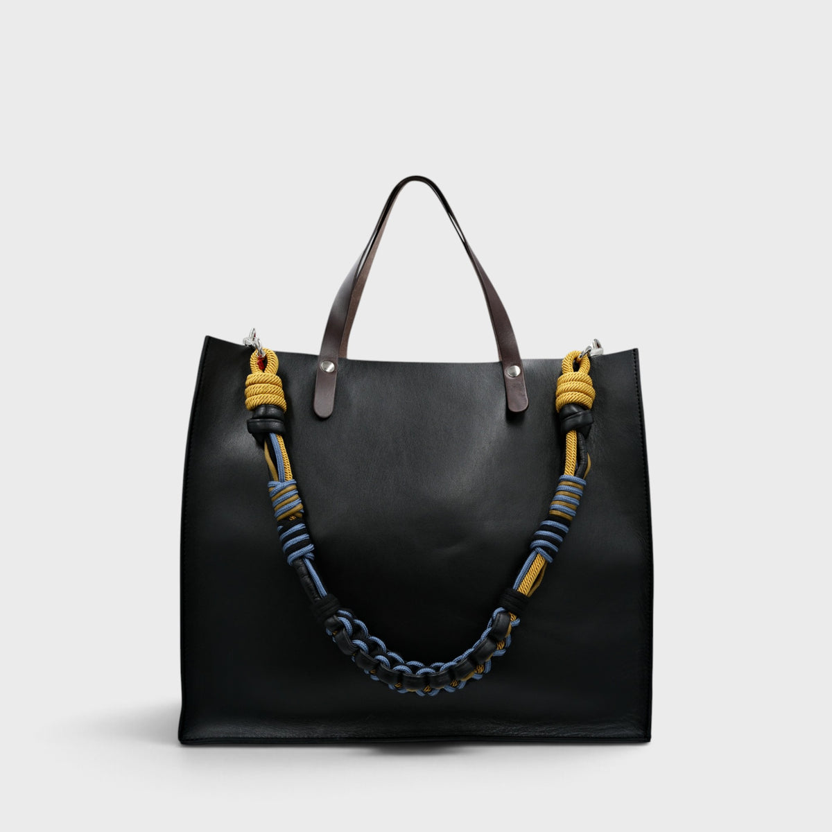 A black leather cabas bag with dark brown leather handles and a distinctive woven shoulder strap featuring yellow, blue, and black stripes, set against a light gray background.