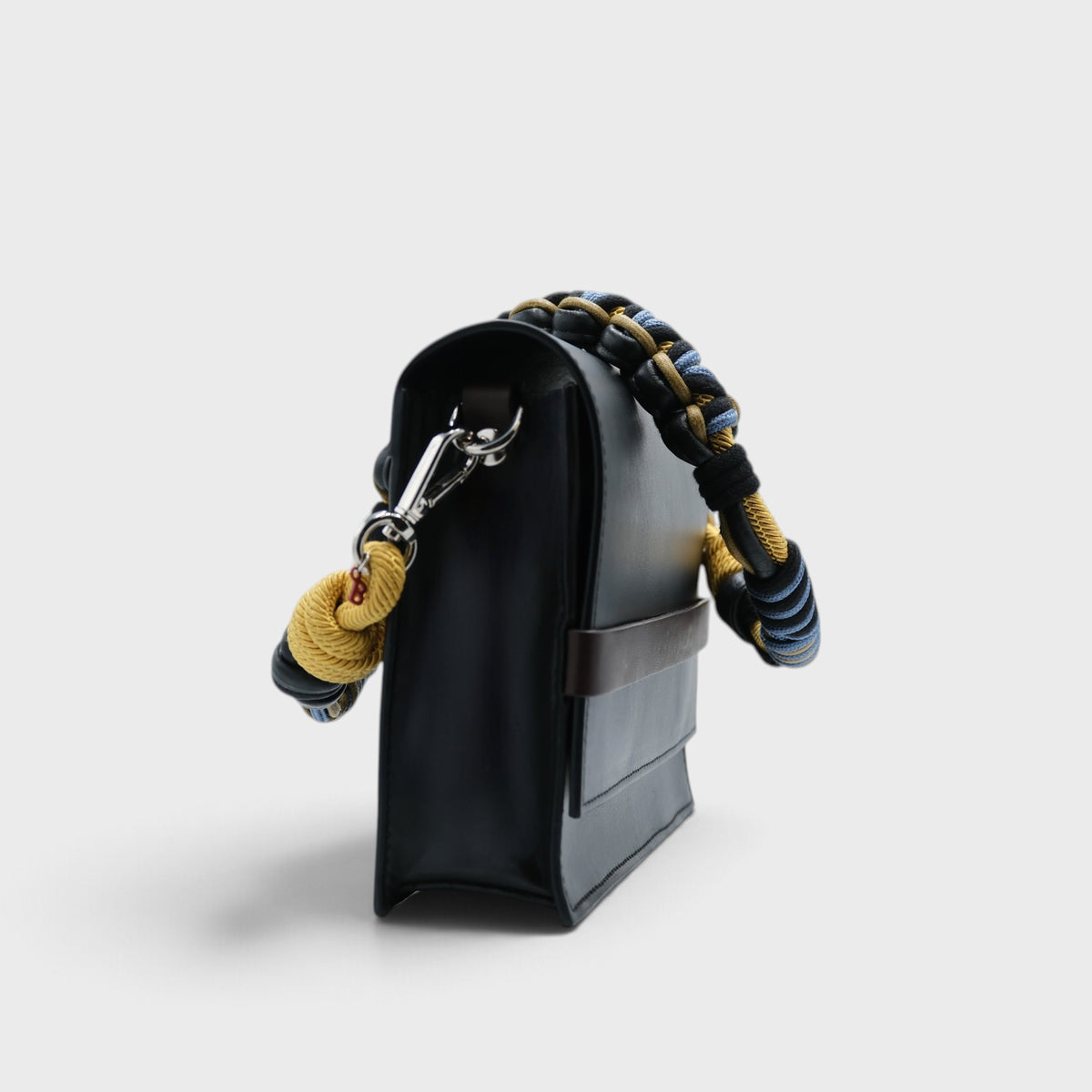A black leather rectangular bag with a colorful macramé strap on a grey background. 