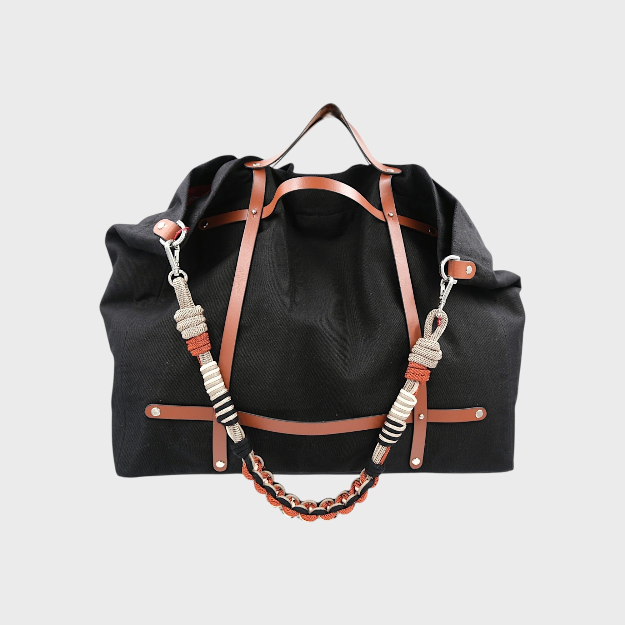 Black canvas weekend or duffle bag with a tan leather harness, paired with a neutral toned shoulder strap in black, white, beige and orange.