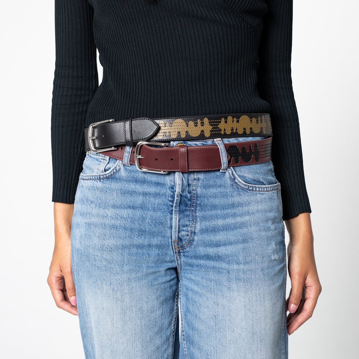 Person wearing a black sweater, blue jeans, and a black leather belt with gold accents and a burgundy leather belt with black accents on a white background
