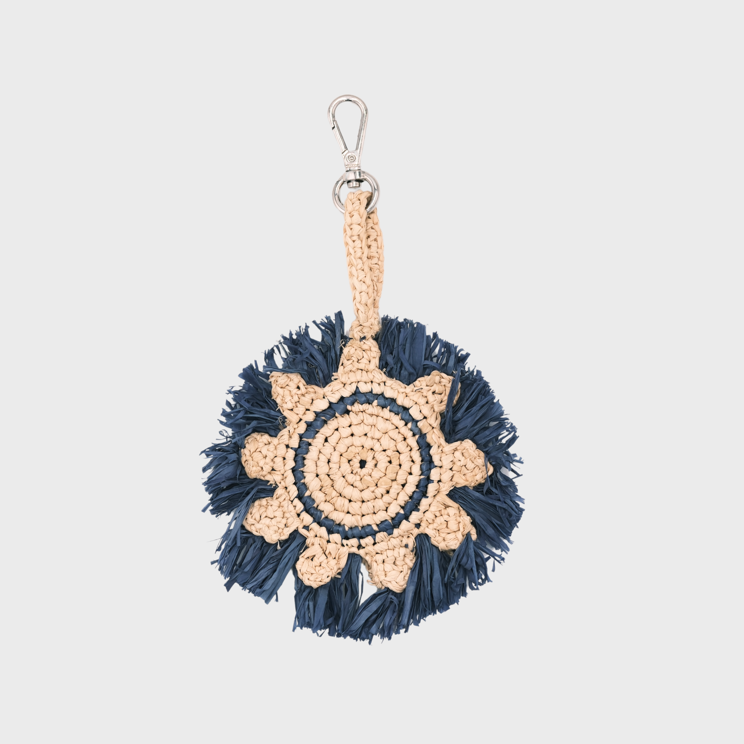 A handmade raffia keychain featuring a circular pattern in natural beige and dark navy blue, representing the sun, finished with a silver metal lobster clasp at the top.