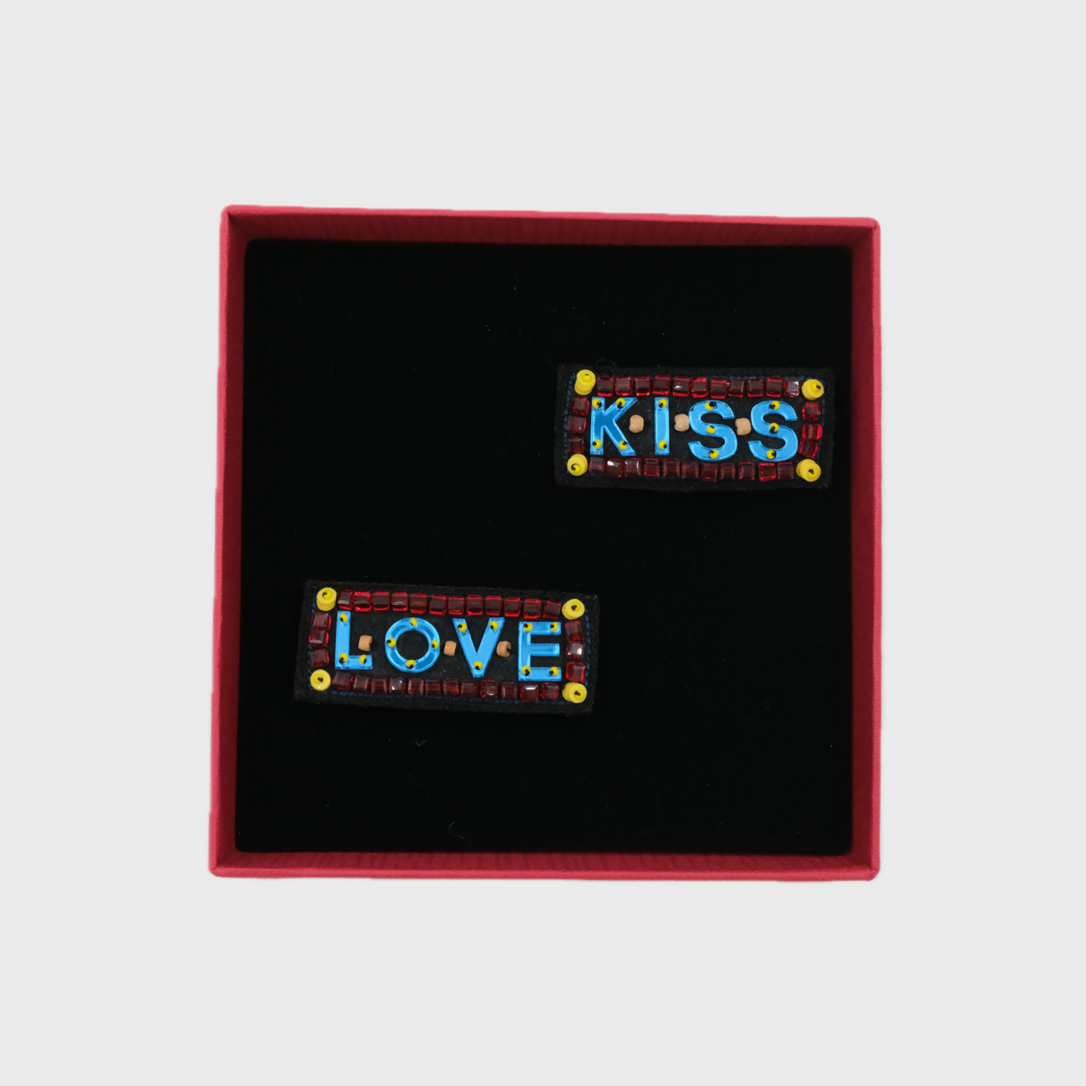 Two embroidered brooches in a red box with written 'kiss' and 'love' on them. 