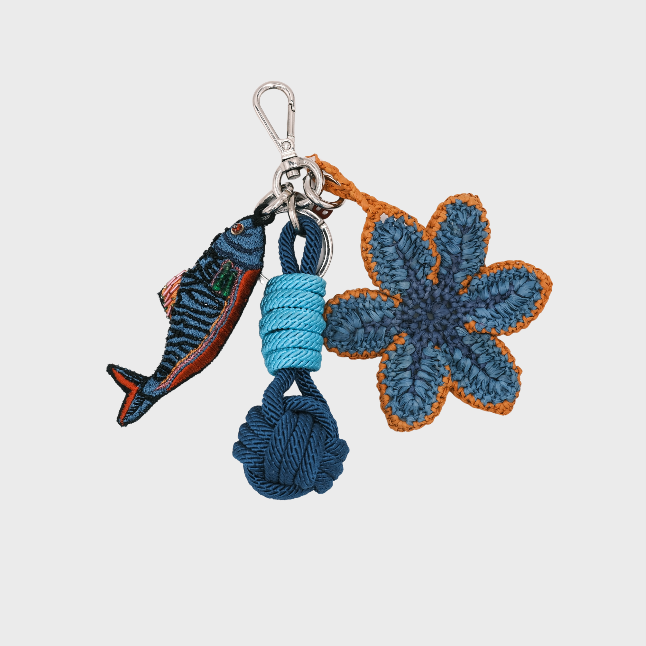A handmade macramé and raffia keychain featuring a crocheted blue flower with orange trim, a blue fish charm, and a light and dark blue knotted ball, attached to a silver clasp.