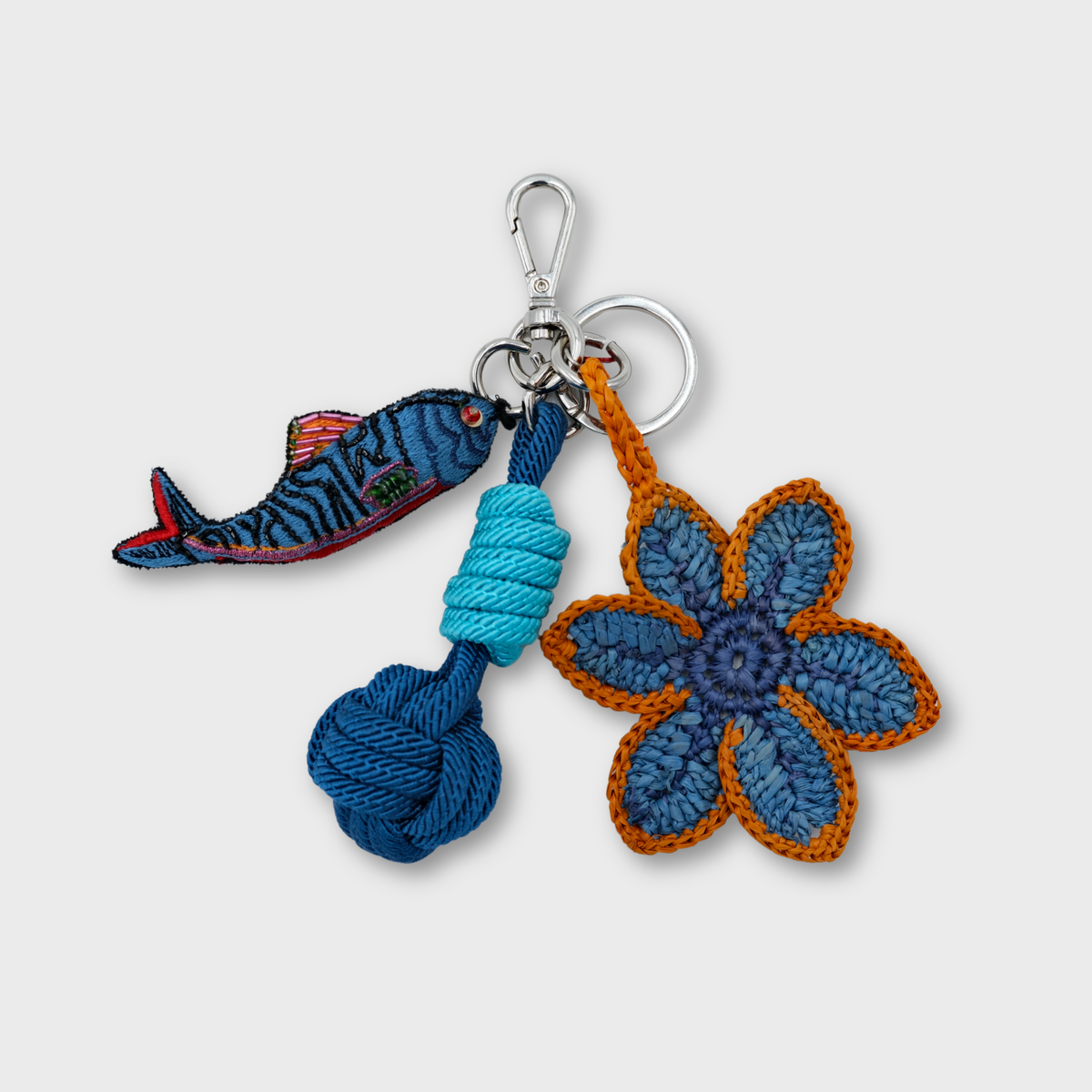 A handmade macramé and raffia keychain featuring a crocheted blue flower with orange trim, a blue fish charm, and a light and dark blue knotted ball, attached to a silver clasp.