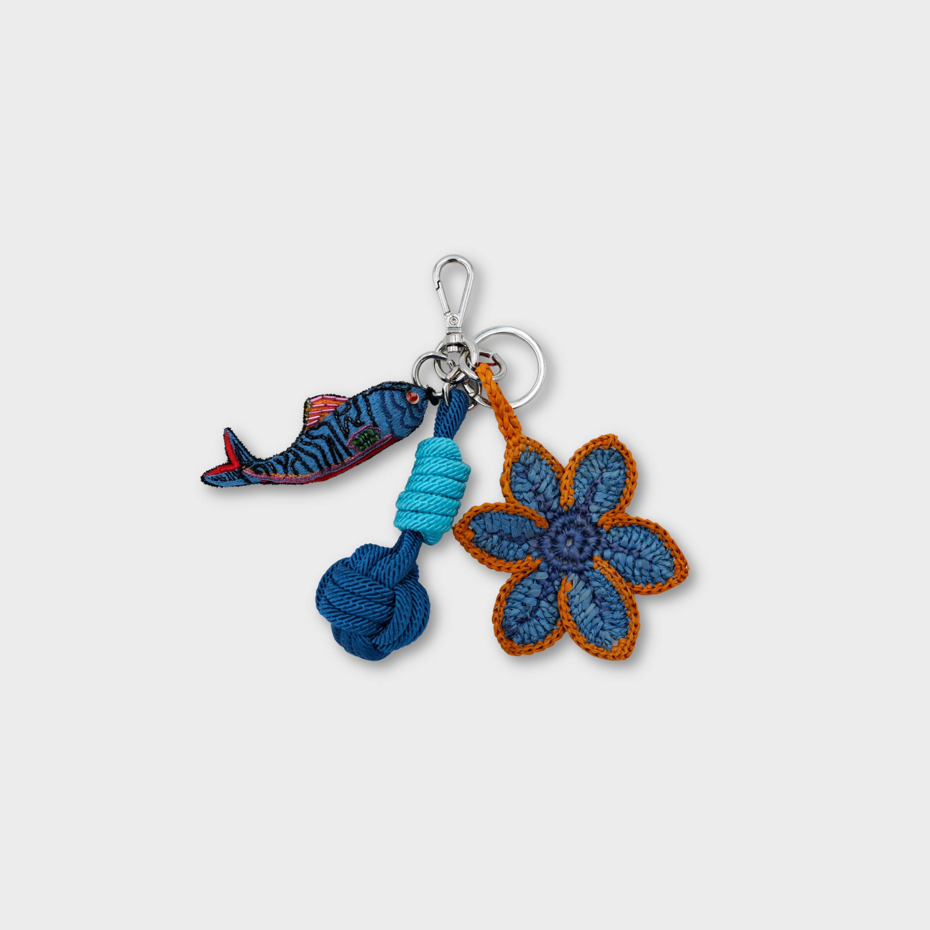 A handmade macramé and raffia keychain featuring a crocheted blue flower with orange trim, a blue fish charm, and a light and dark blue knotted ball, attached to a silver clasp.