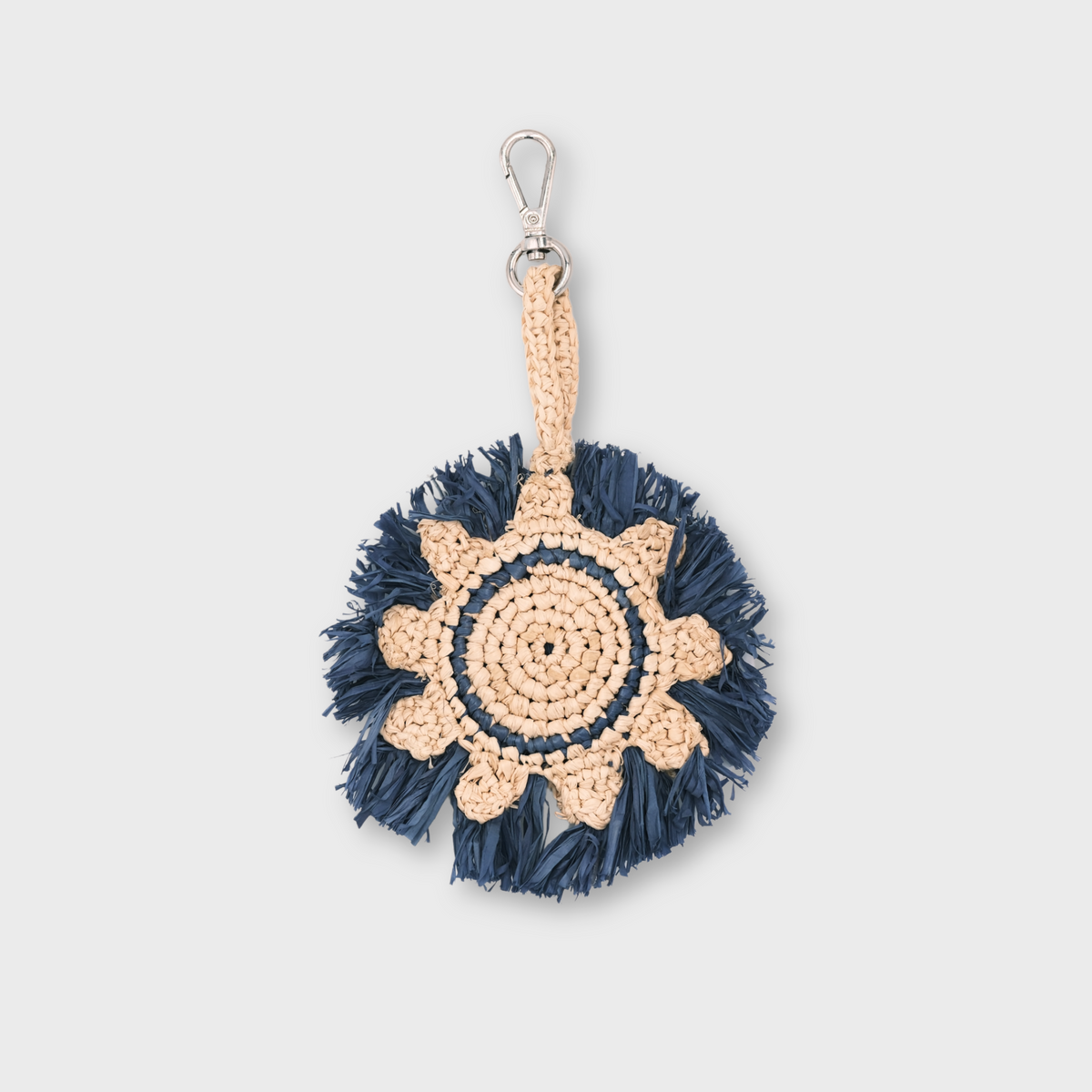 A handmade raffia keychain featuring a circular pattern in natural beige and dark navy blue, representing the sun, finished with a silver metal lobster clasp at the top.
