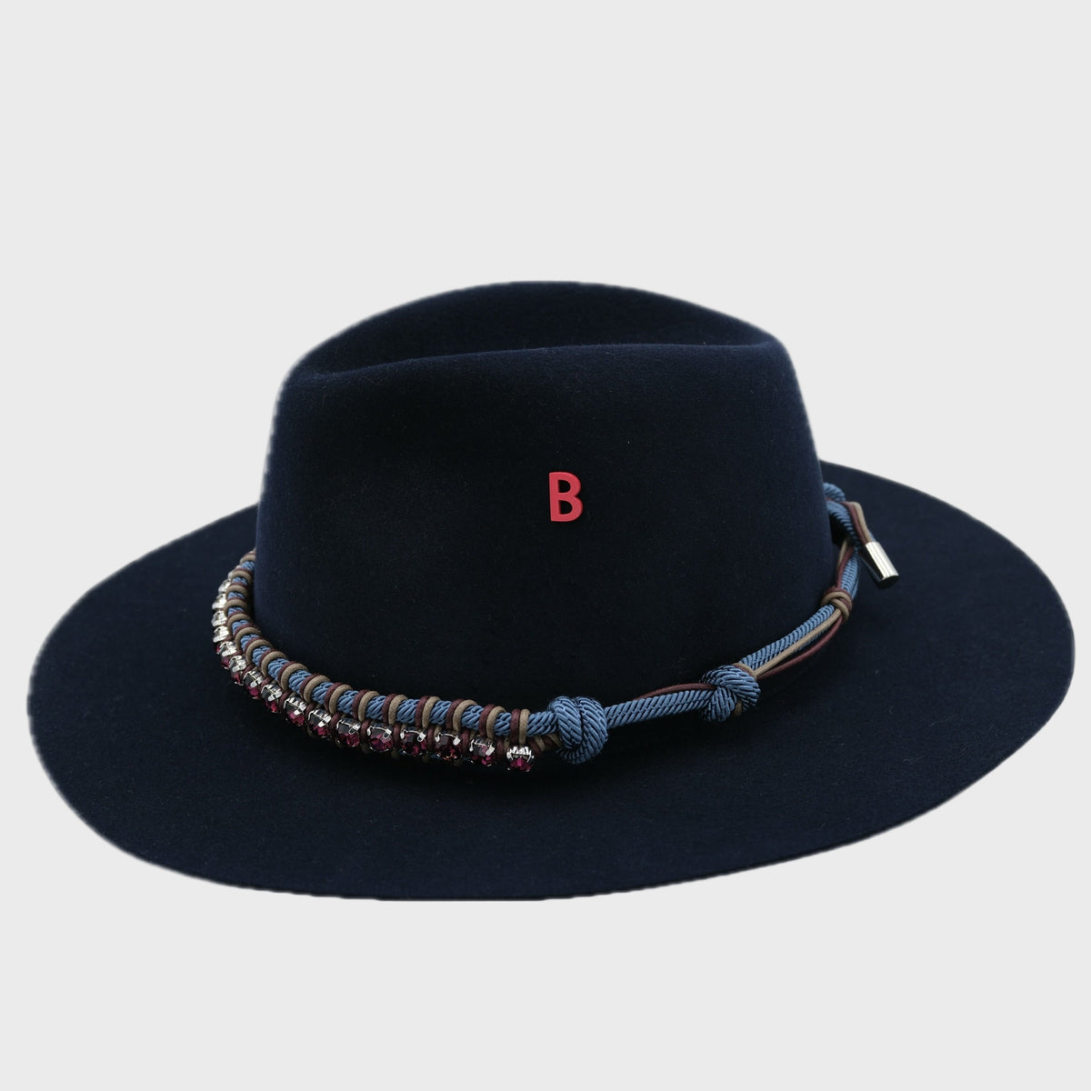 Black wool fedora hat with decorative band woth blue and purple macramé and purple rhinestones, and red letter 'B' înned on it on a light gray background