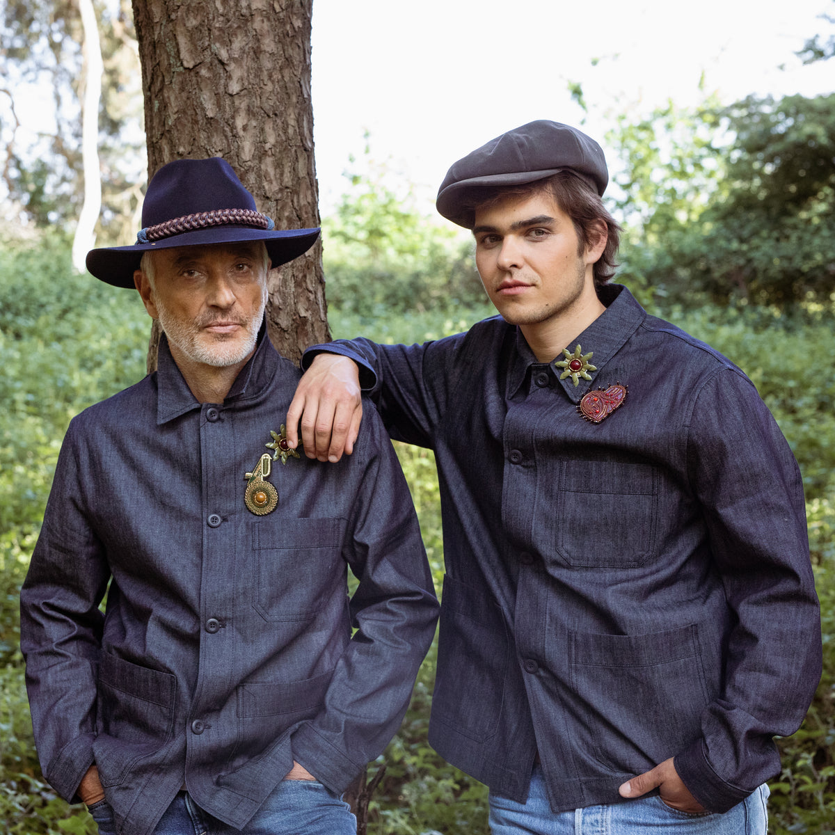 Two men in matching dark jackets and one with a blue hat with a macrame band standing in a forest setting.