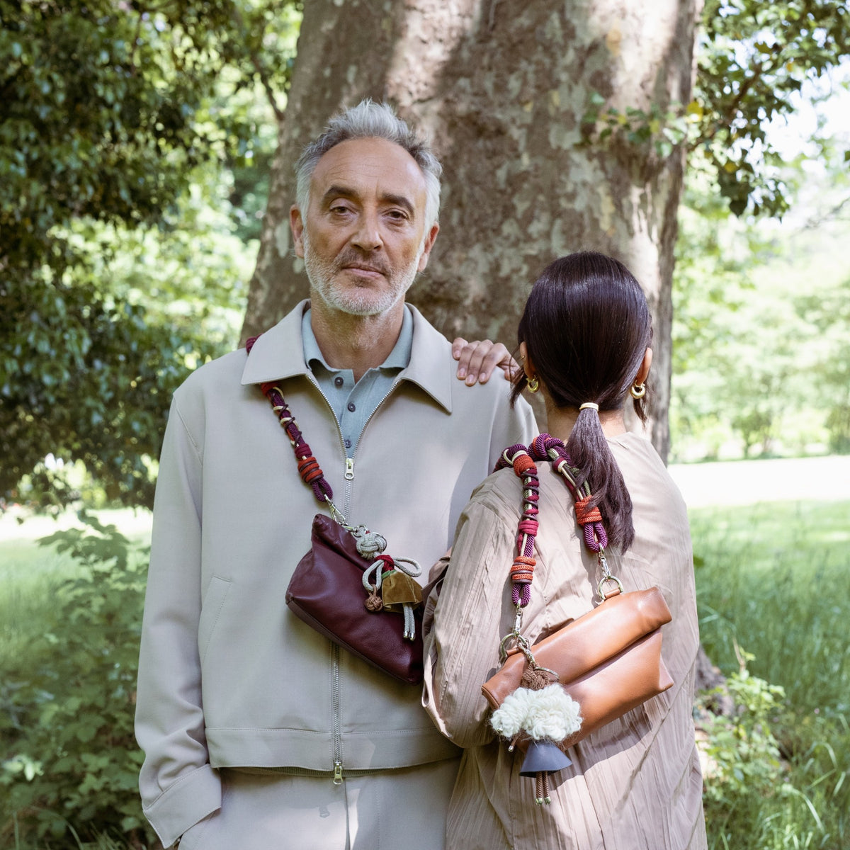 Two people standing outdoors with leather pouch, surrounded by trees and greenery.