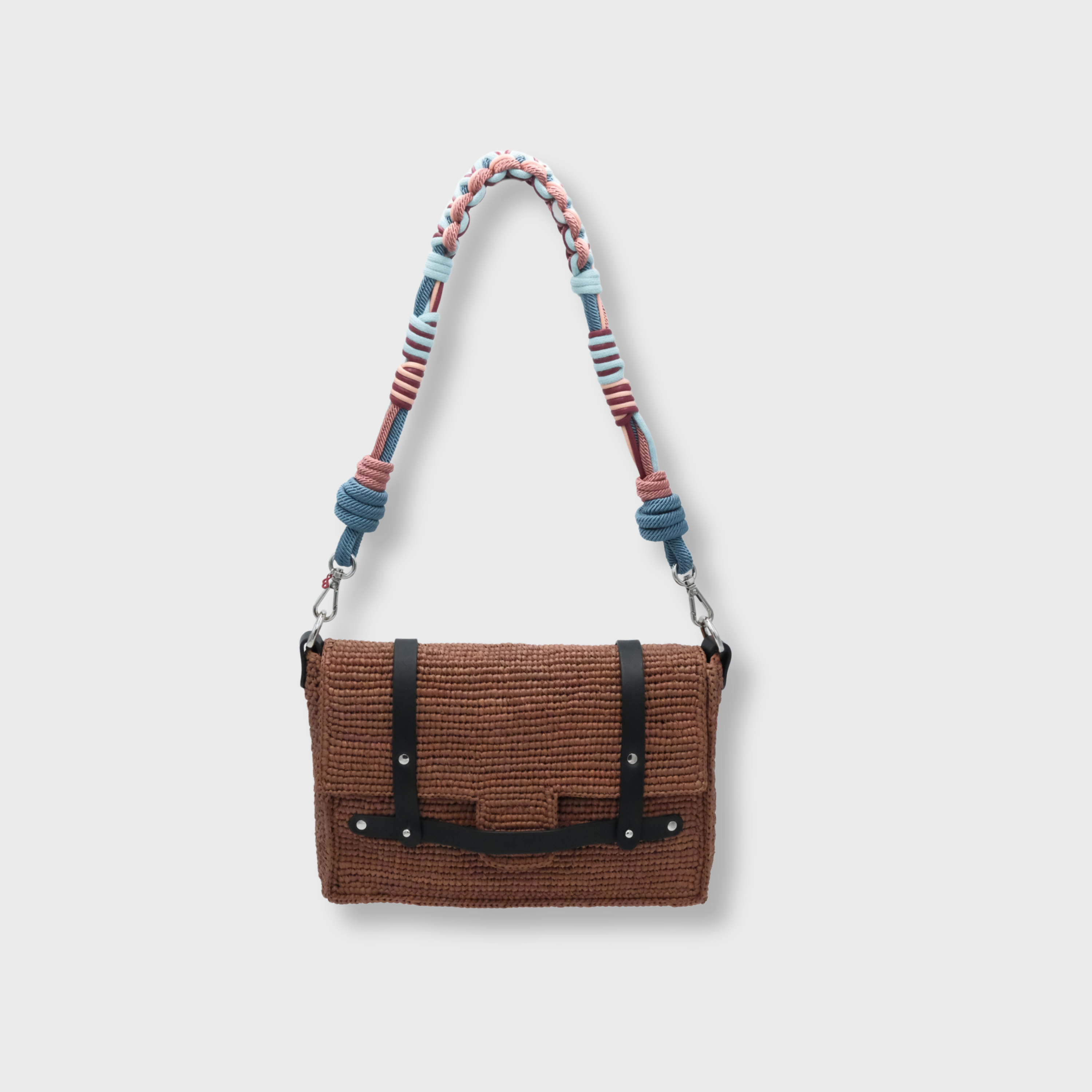 A brown raffia shoulder bag with black leather straps hanging by a colorful macramé braided strap featuring pink, blue, set against a light gray background.