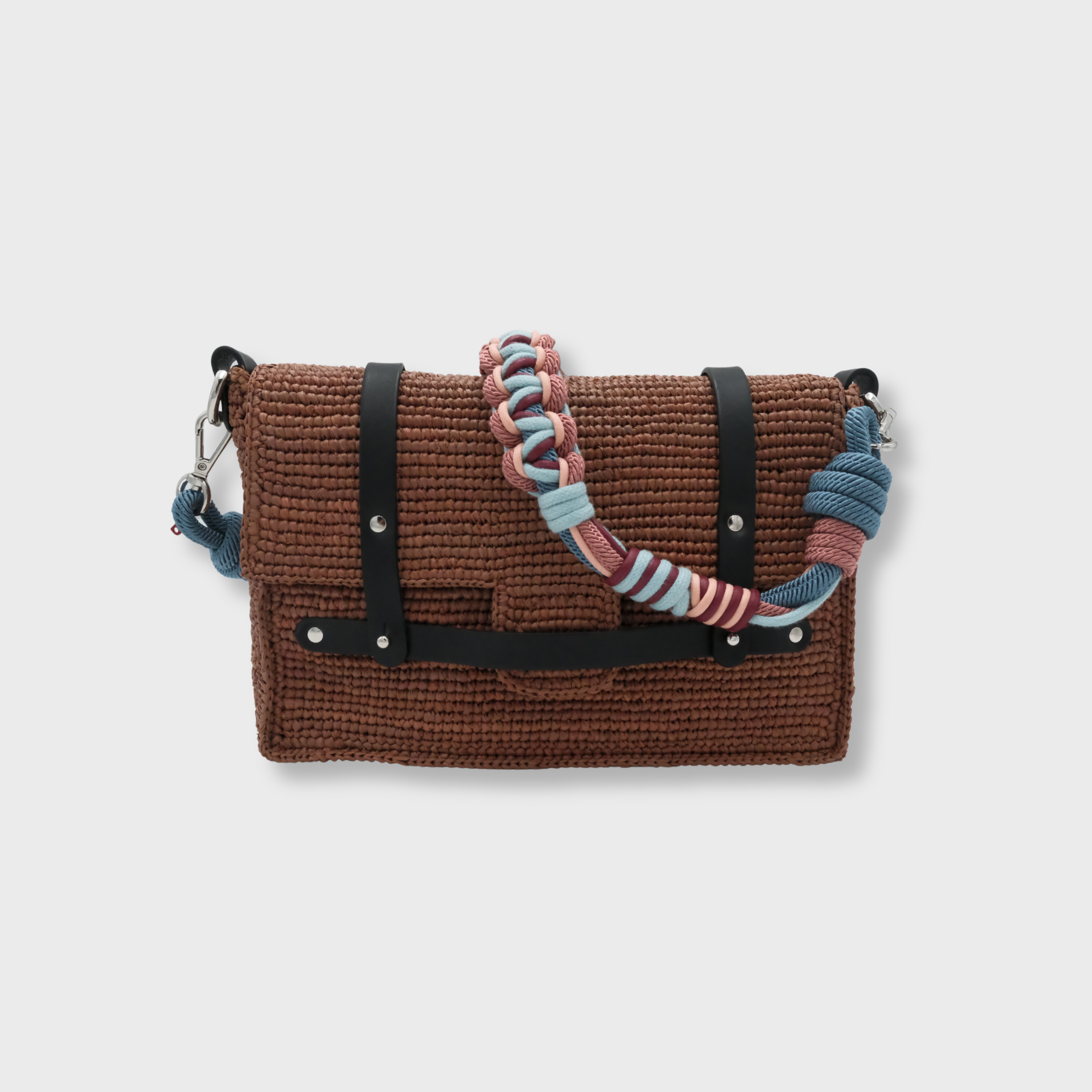 A brown raffia shoulder bag with black leather straps and a colorful macramé braided strap featuring pink and blue. 