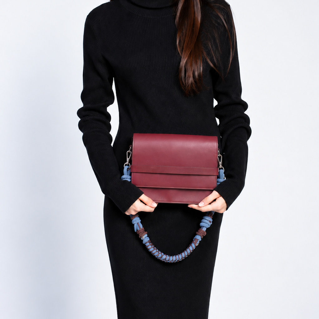 A woman wearing a black turtleneck sweater and black skirt holds a burgundy leather clutch bag with a blue braided strap against a light gray studio background.