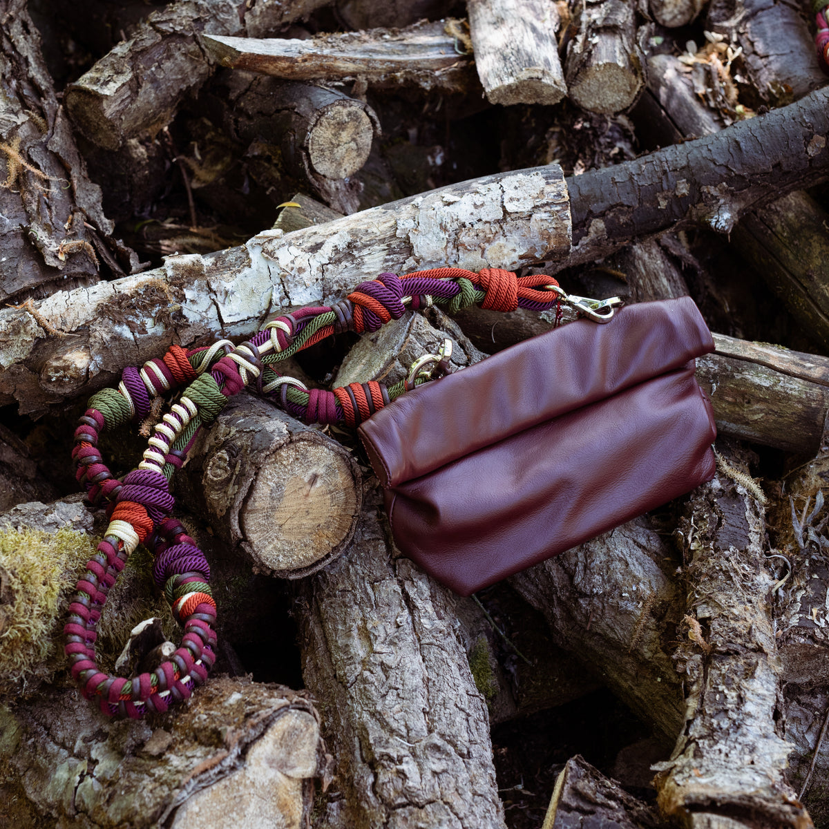 A burgundy leather pouch with a burgundy macramé woven strap, in a forrest. 