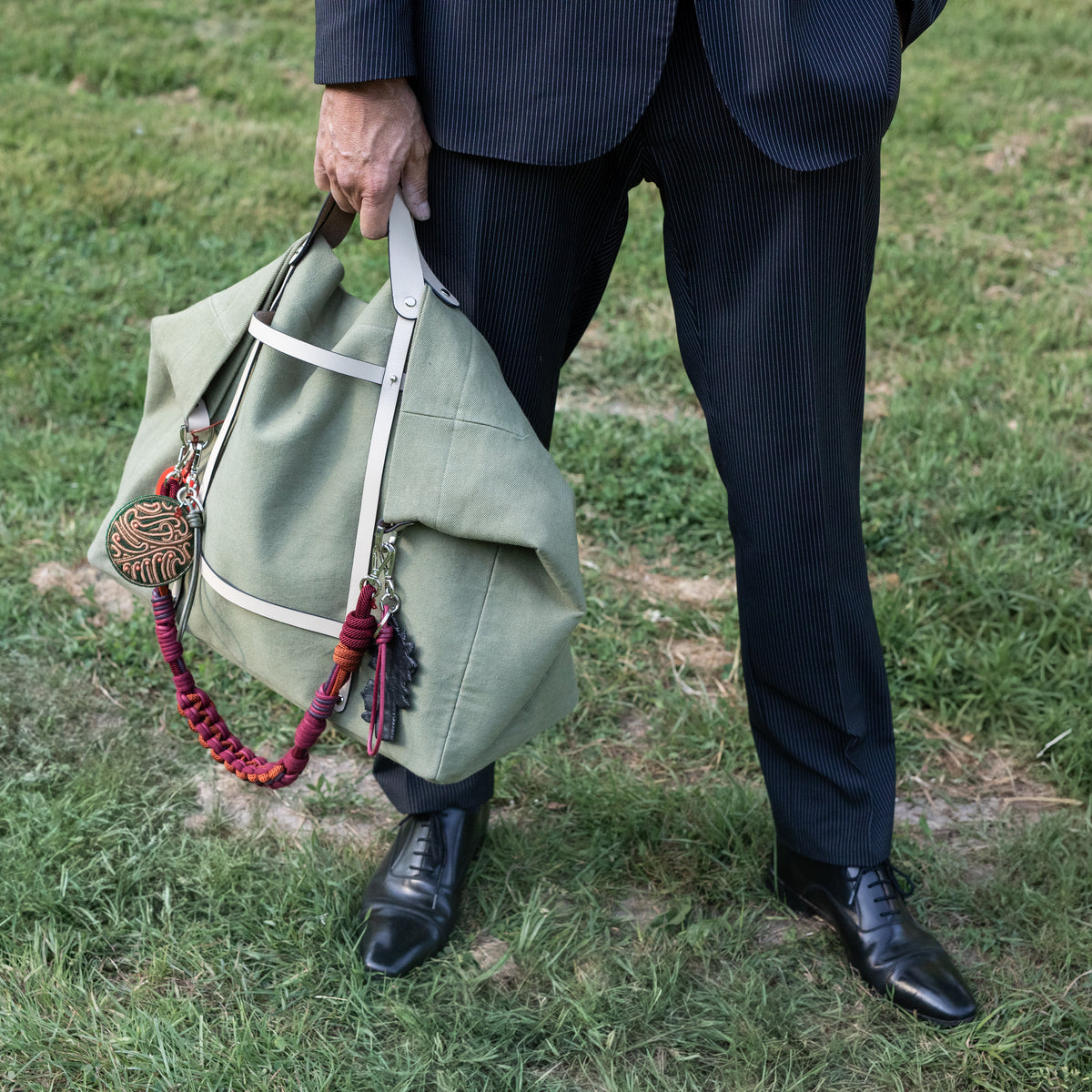 Person holding a green bag with a burgundy decorative macramé strap on grass